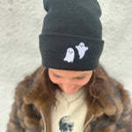 Woman wearing Black beanie with white embroidered ghosts on the folded brim for Halloween, wearing a cream sweater with black skulls and a brown faux fur jacket with silver zipper