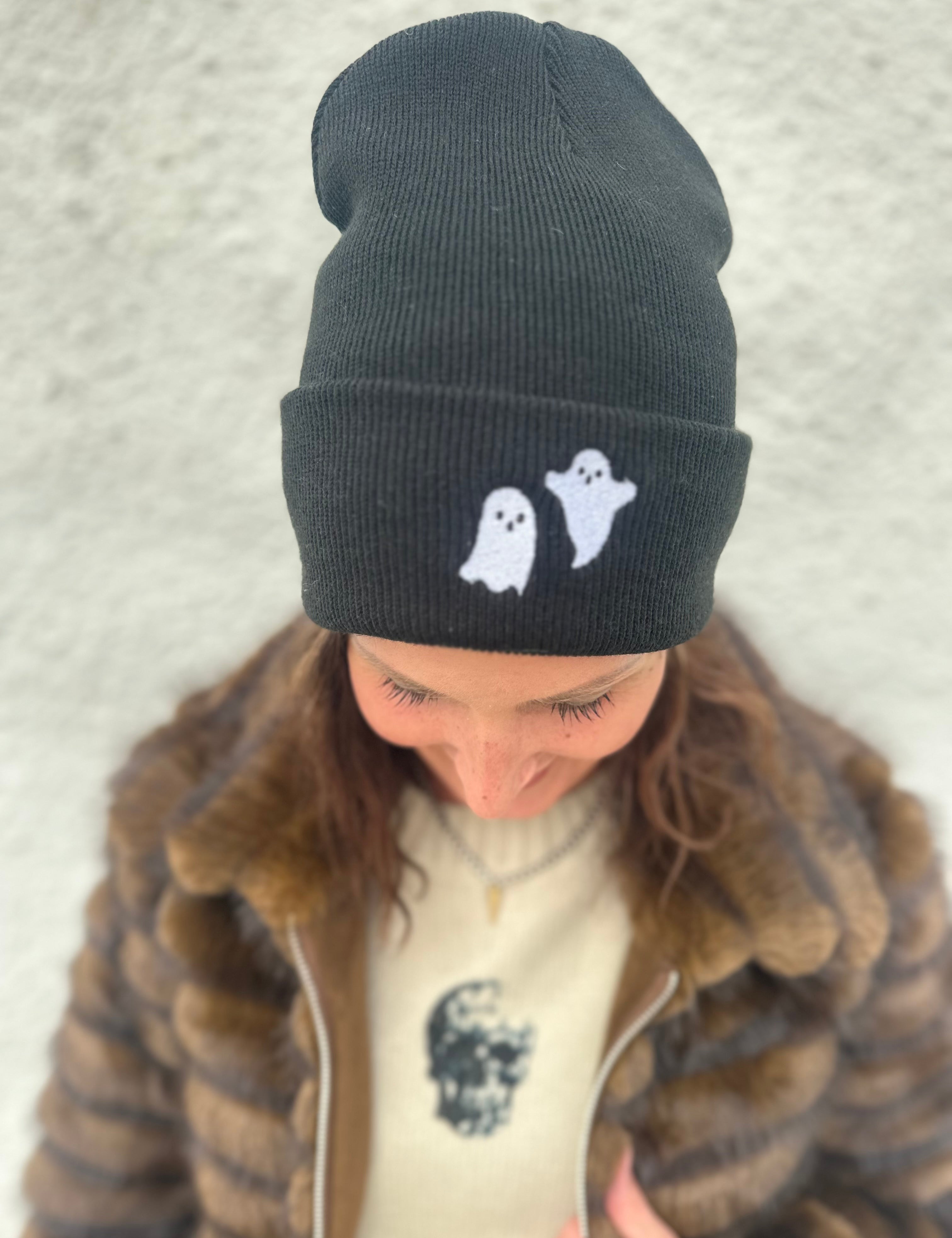 Woman wearing Black beanie with white embroidered ghosts on the folded brim for Halloween, wearing a cream sweater with black skulls and a brown faux fur jacket with silver zipper