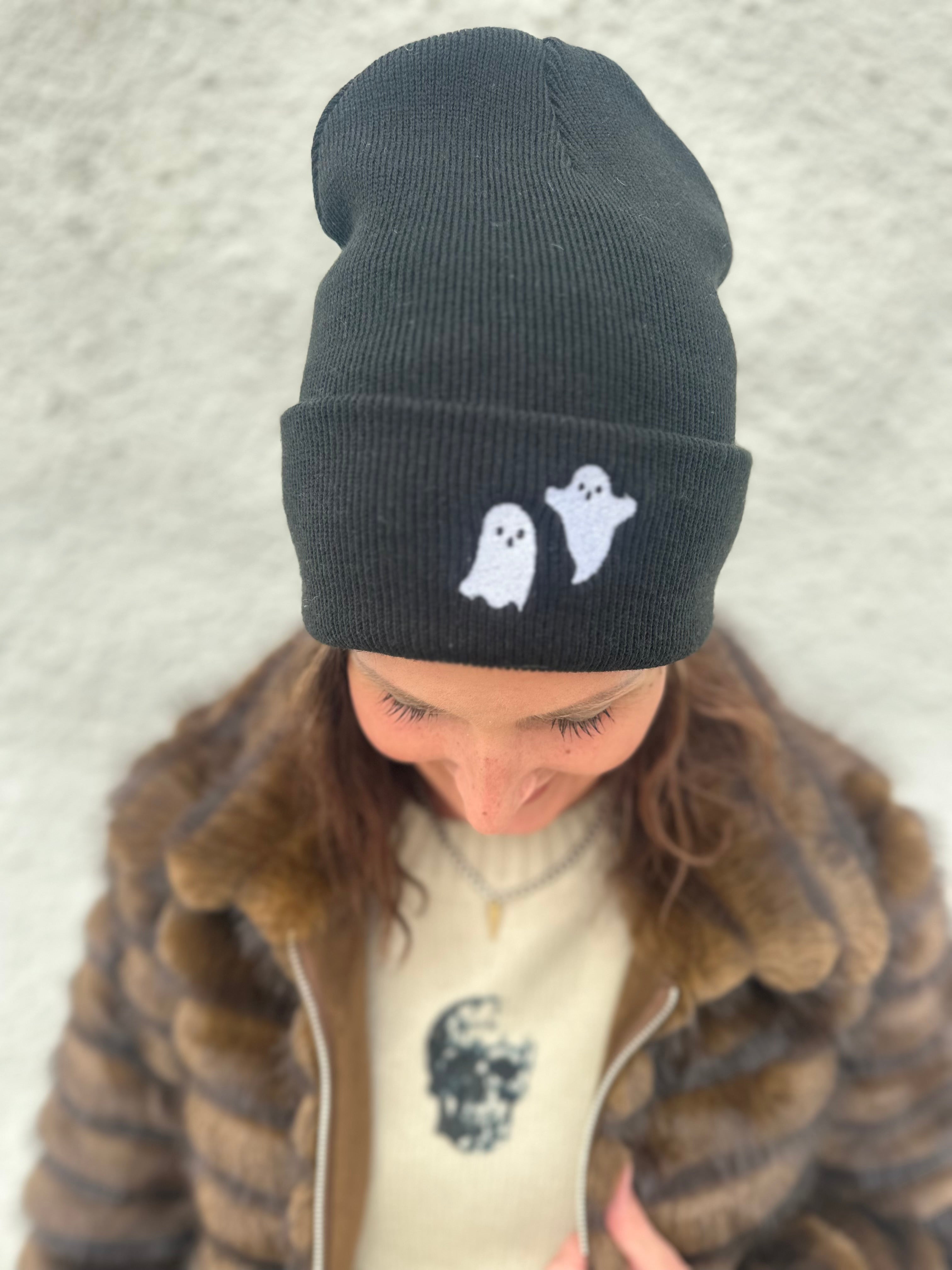 Woman wearing Black beanie with white embroidered ghosts on the folded brim for Halloween, wearing a cream sweater with black skulls and a brown faux fur jacket with silver zipper