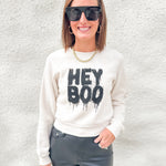 A woman wearing a cream-colored crewneck sweatshirt with a black 'HEY BOO' graphic, paired with dark gray pants, sunglasses, and a gold chain necklace, standing against a textured white wall.