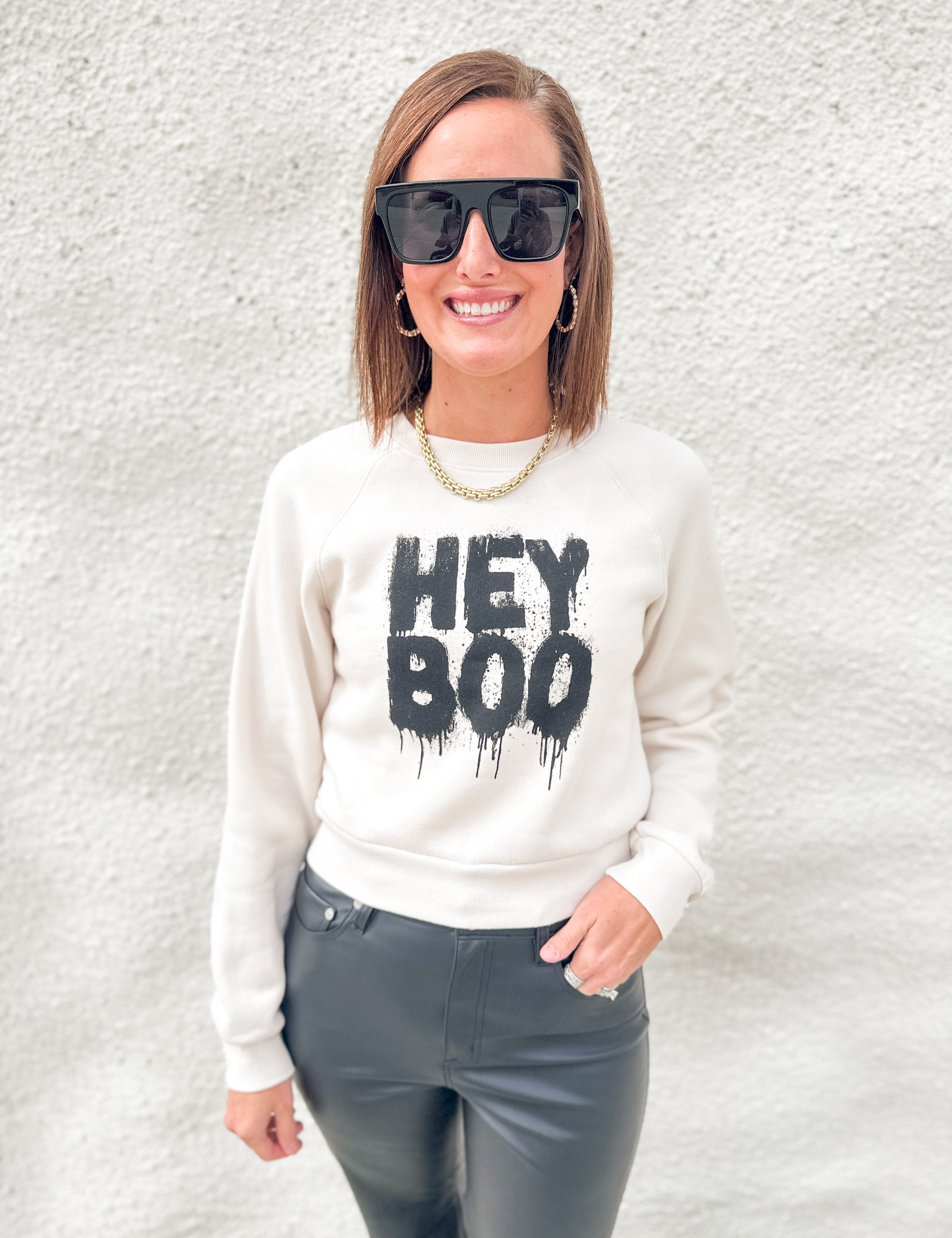 A woman wearing a cream-colored crewneck sweatshirt with a black 'HEY BOO' graphic, paired with dark gray pants, sunglasses, and a gold chain necklace, standing against a textured white wall.