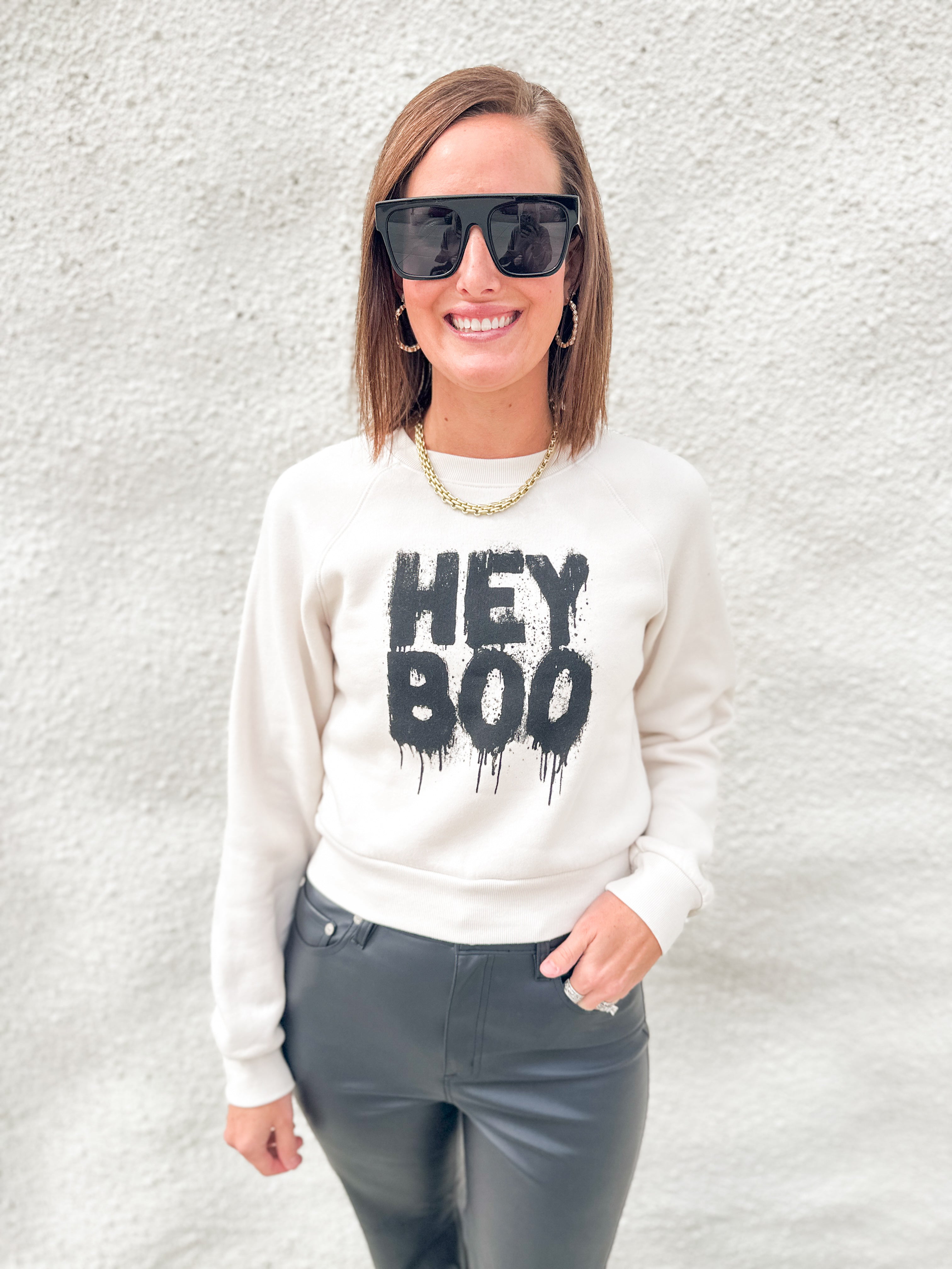 A woman wearing a cream-colored crewneck sweatshirt with a black 'HEY BOO' graphic, paired with dark gray pants, sunglasses, and a gold chain necklace, standing against a textured white wall.