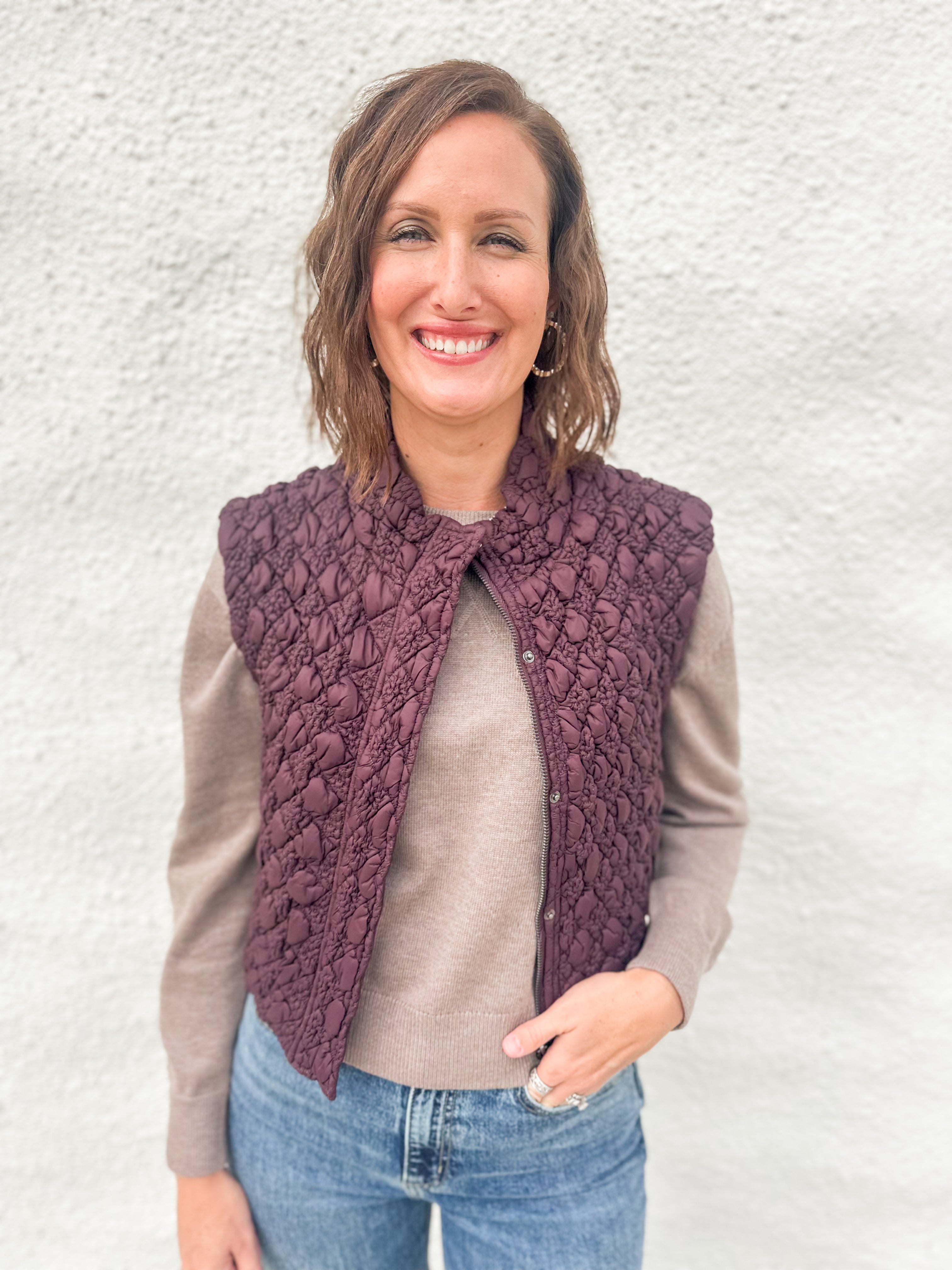 A woman wearing a deep burgundy quilted vest with zipper front over a lightweight wool blend sweater in a mocha color and denim jeans from Daze
