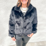 A woman wearing a dark gray faux fur jacket, black sunglasses, a white t-shirt, and black leather pants, standing against a textured white wall.