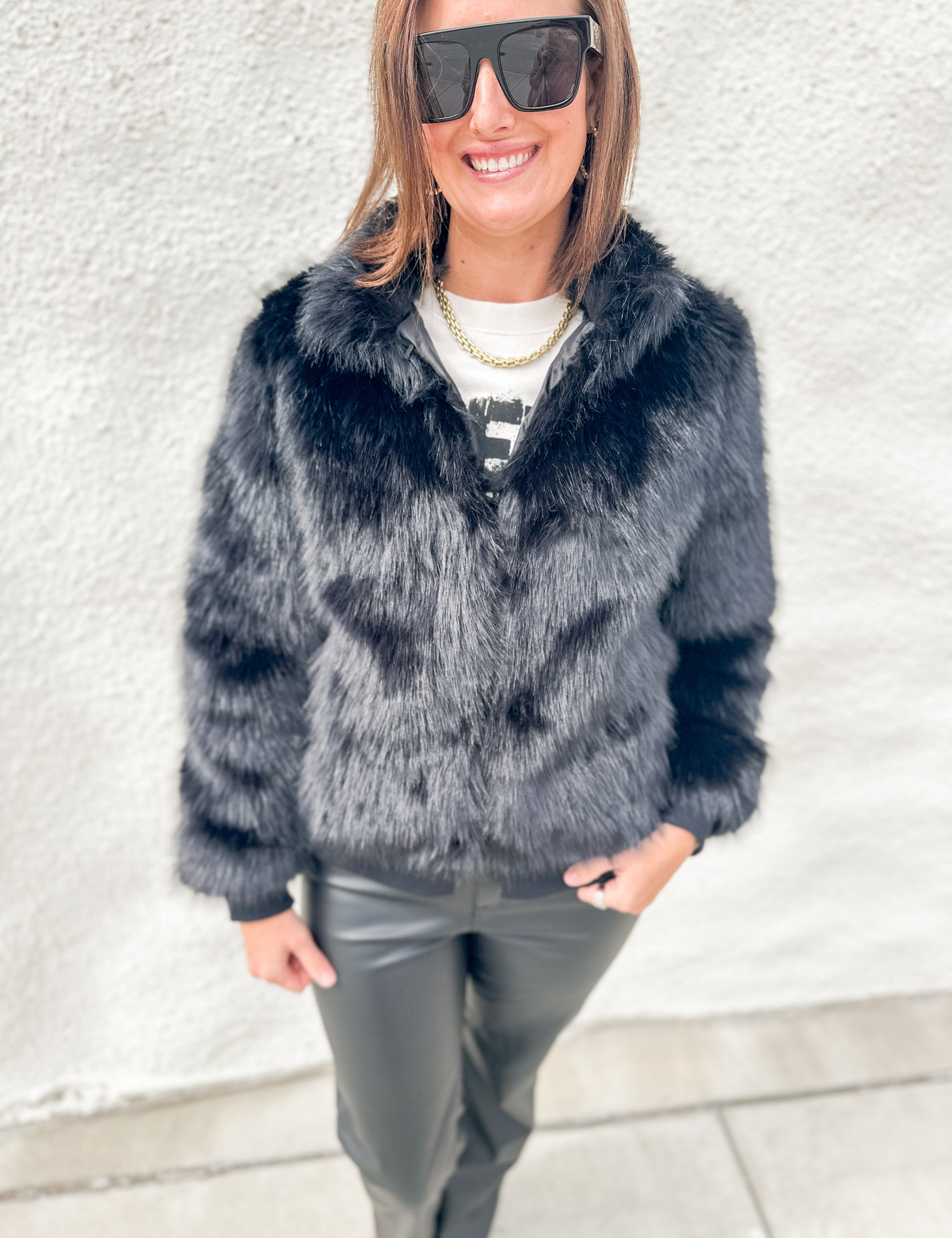 A woman wearing a dark gray faux fur jacket, black sunglasses, a white t-shirt, and black leather pants, standing against a textured white wall.
