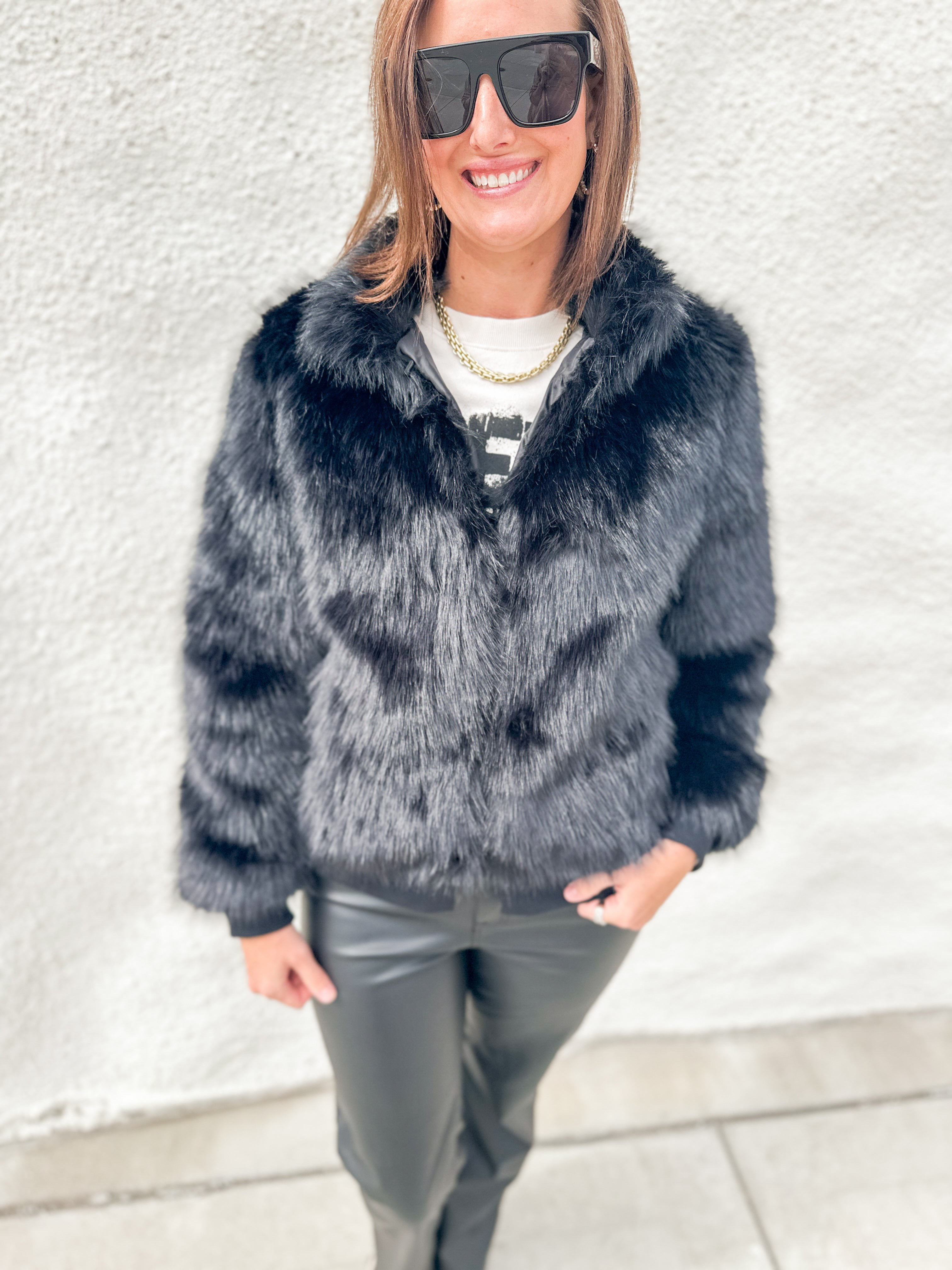 A woman wearing a dark gray faux fur jacket, black sunglasses, a white t-shirt, and black leather pants, standing against a textured white wall.