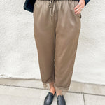 A person wearing loose taupe-brown drawstring pants with sheer lace trim at the ankles, styled with a dark navy knit sweater and black loafers against a white stucco wall.