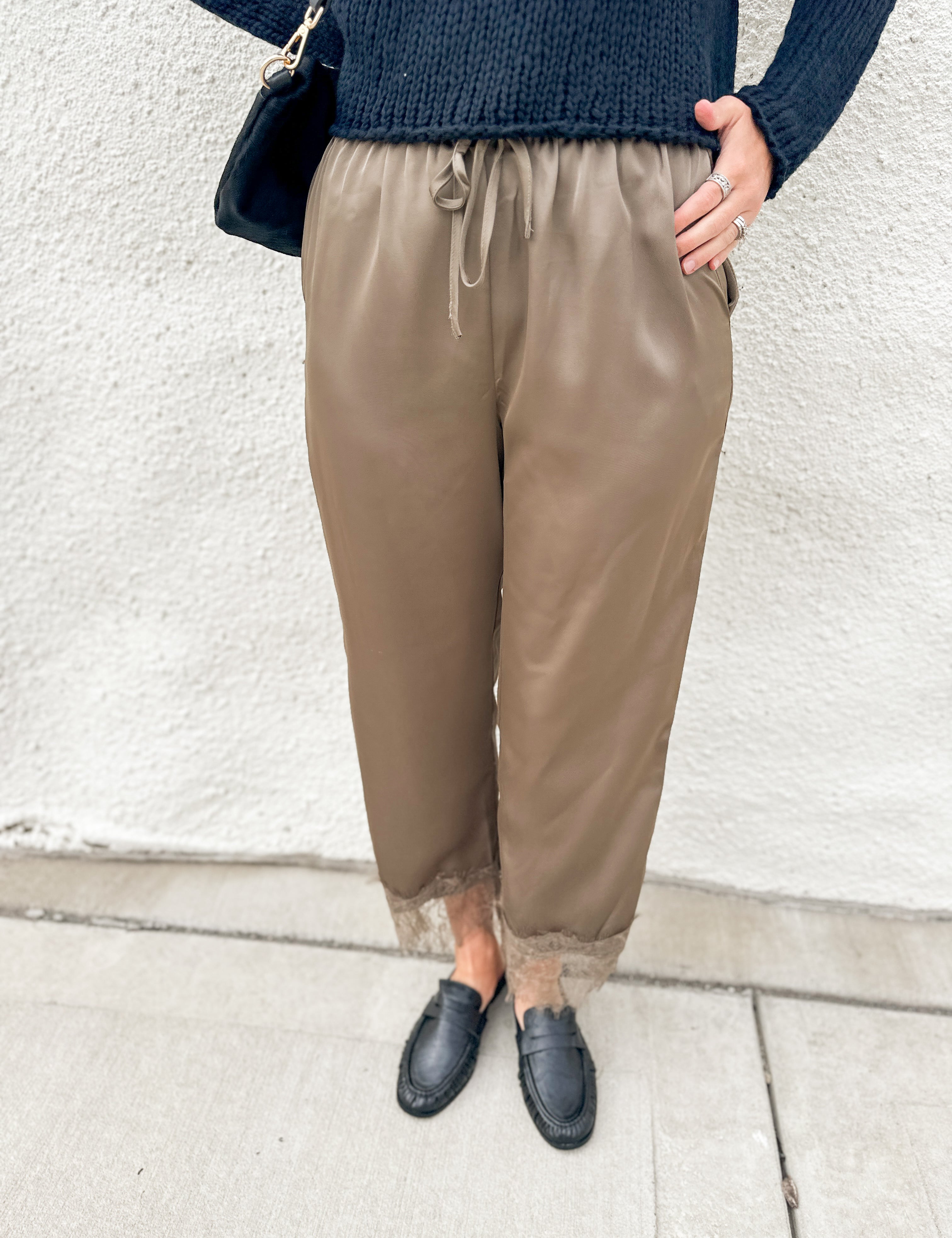 A person wearing loose taupe-brown drawstring pants with sheer lace trim at the ankles, styled with a dark navy knit sweater and black loafers against a white stucco wall.