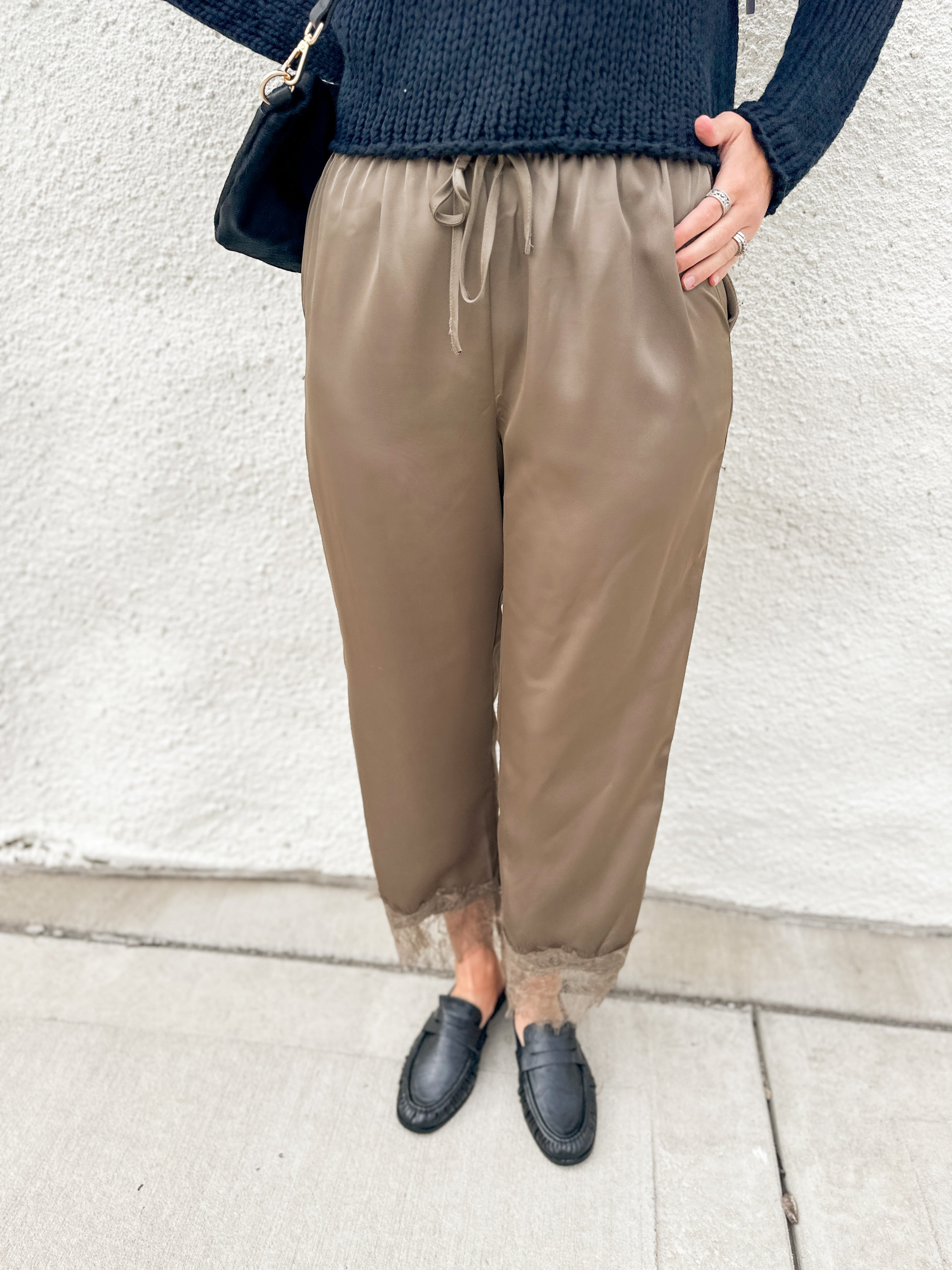 A person wearing loose taupe-brown drawstring pants with sheer lace trim at the ankles, styled with a dark navy knit sweater and black loafers against a white stucco wall.