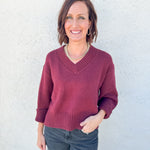 A woman wearing a burgundy v-neck knit sweater and dark gray jeans, standing against a textured white stucco wall.