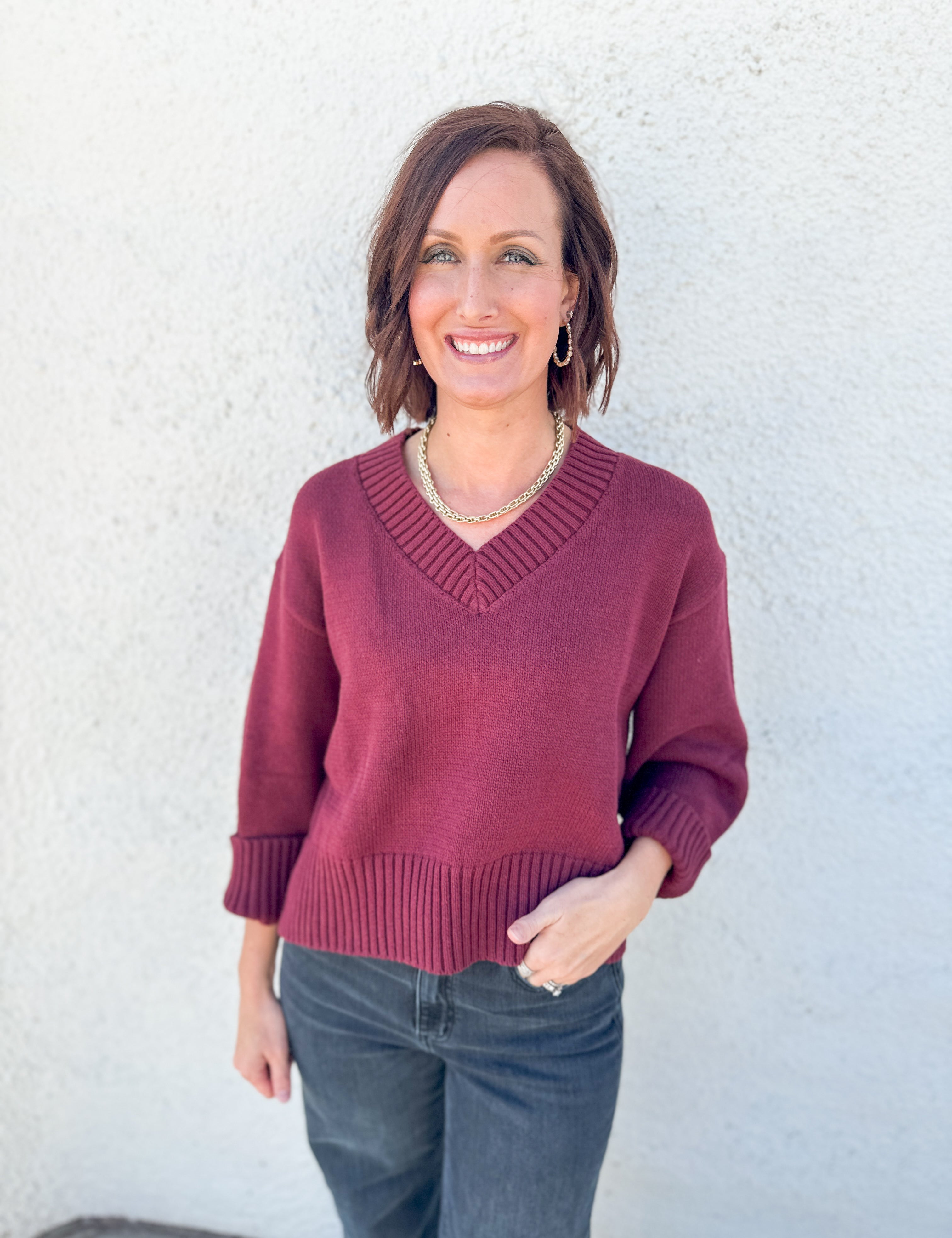 A woman wearing a burgundy v-neck knit sweater and dark gray jeans, standing against a textured white stucco wall.