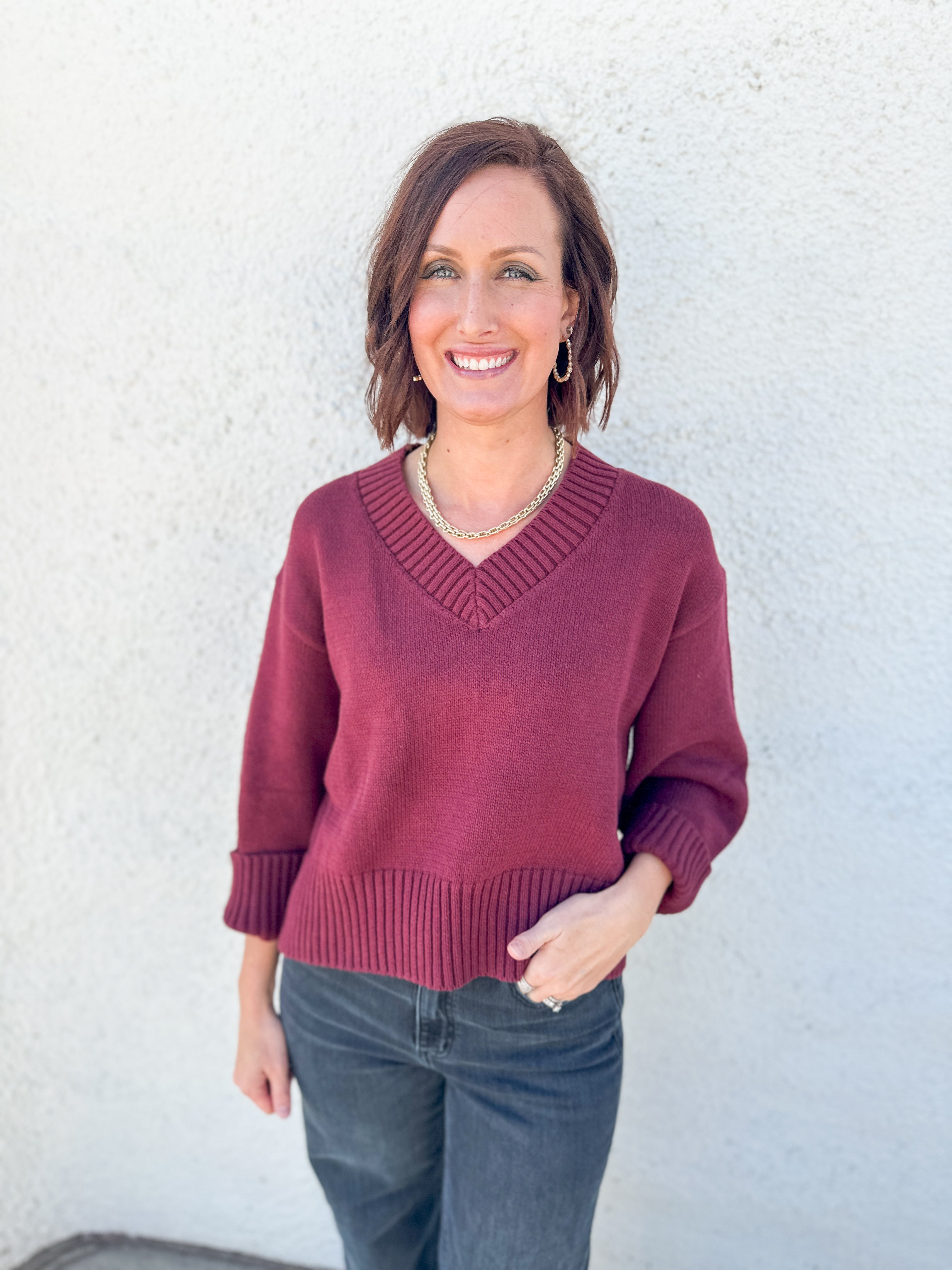 A woman wearing a burgundy v-neck knit sweater and dark gray jeans, standing against a textured white stucco wall.