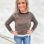 A woman with blonde hair wearing a brown long-sleeve crewneck sweater with brown and white striped cuffs and hem, paired with blue jeans, standing against a white textured wall.