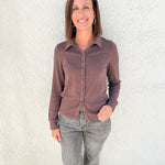 A woman wearing a long-sleeve button-up shirt in a muted brownish-purple color and gray jeans, standing against a white stucco wall.