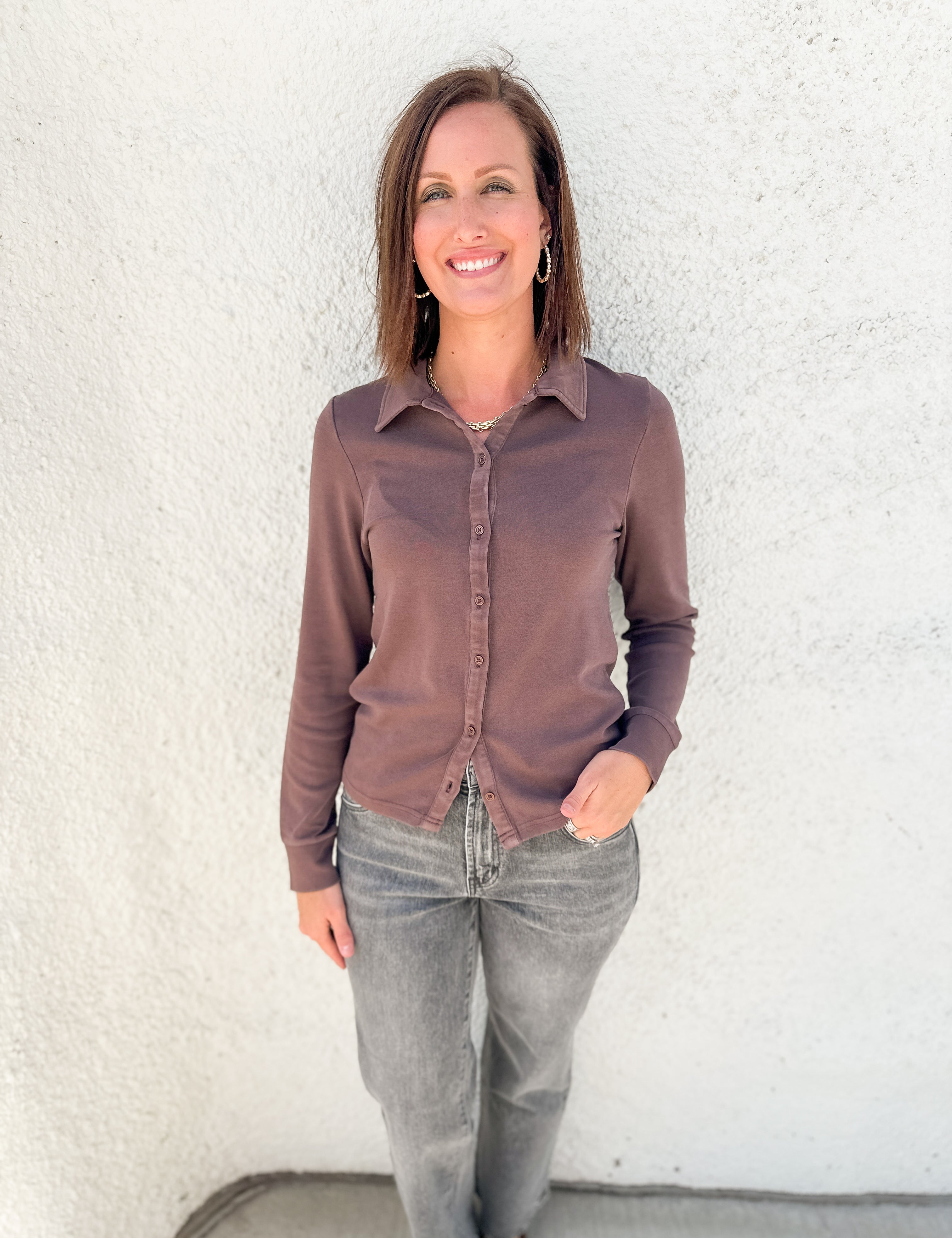 A woman wearing a long-sleeve button-up shirt in a muted brownish-purple color and gray jeans, standing against a white stucco wall.
