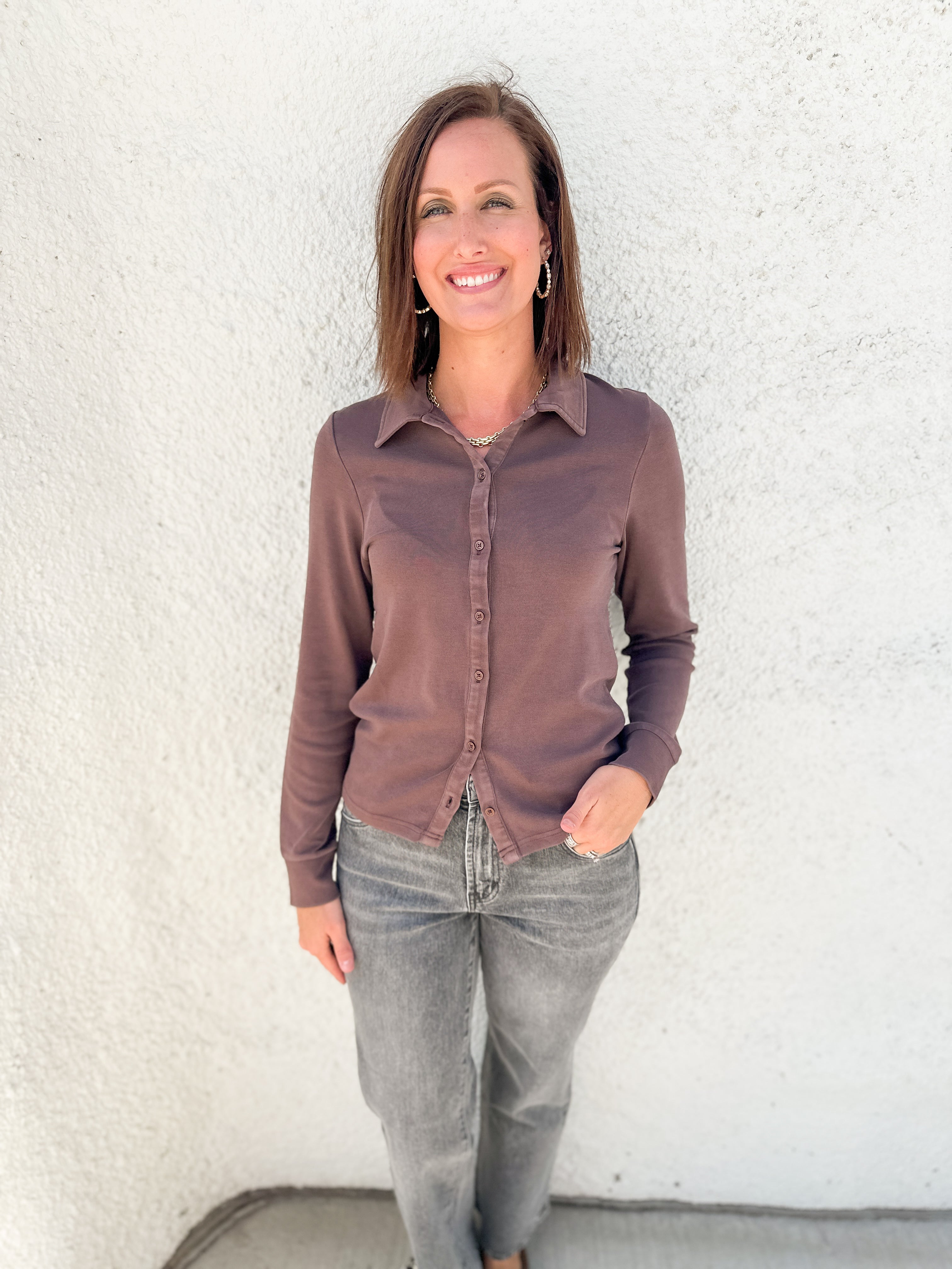 A woman wearing a long-sleeve button-up shirt in a muted brownish-purple color and gray jeans, standing against a white stucco wall.