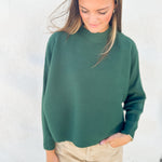 A woman wearing a dark green ribbed knit mock neck sweater and beige pants, standing against a light textured wall.
