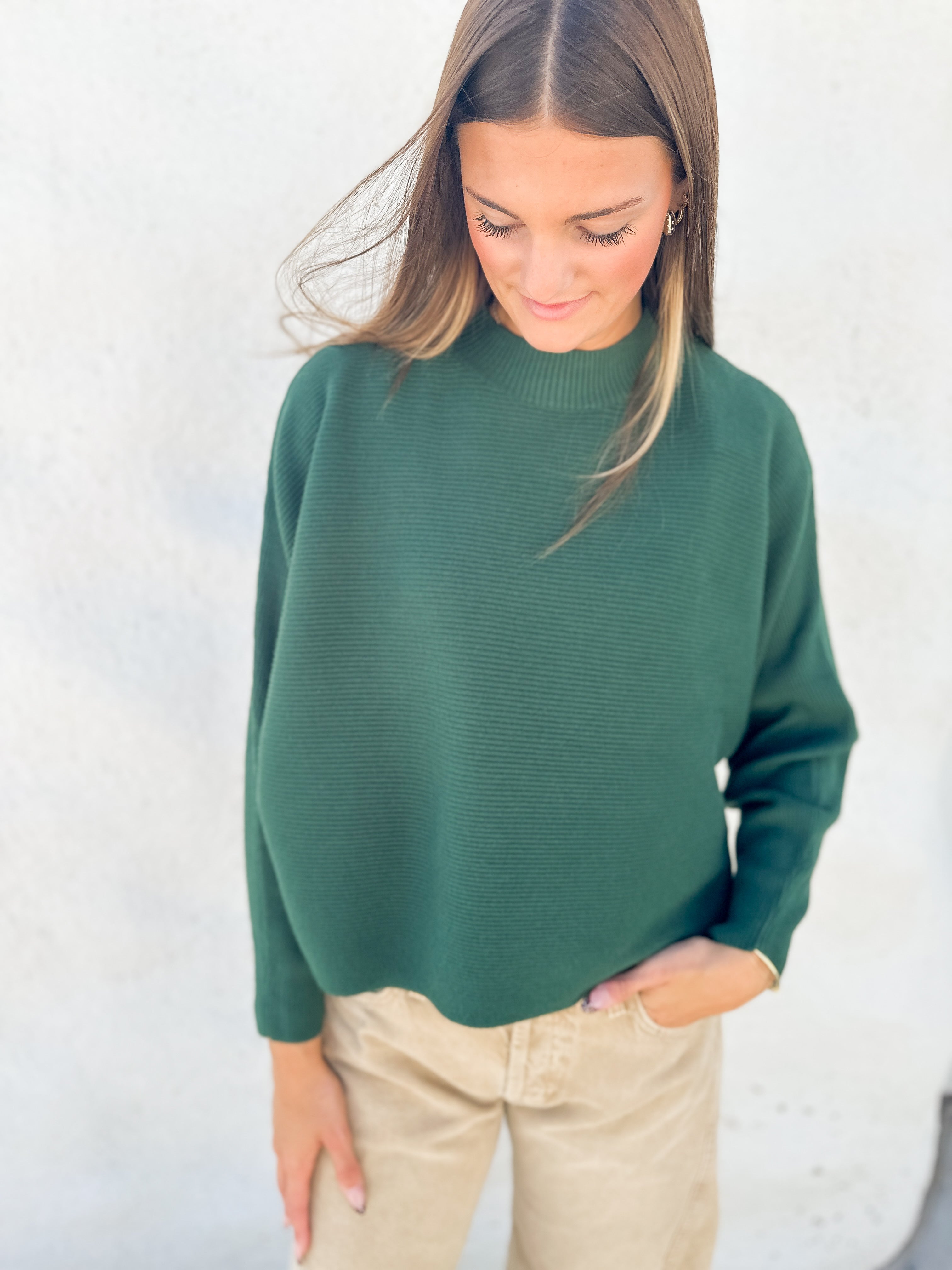 A woman wearing a dark green ribbed knit mock neck sweater and beige pants, standing against a light textured wall.
