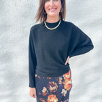 A woman modeling a black ribbed long-sleeve top and a dark floral skirt with orange, purple, and cream flowers, standing against a light gray textured wall.
