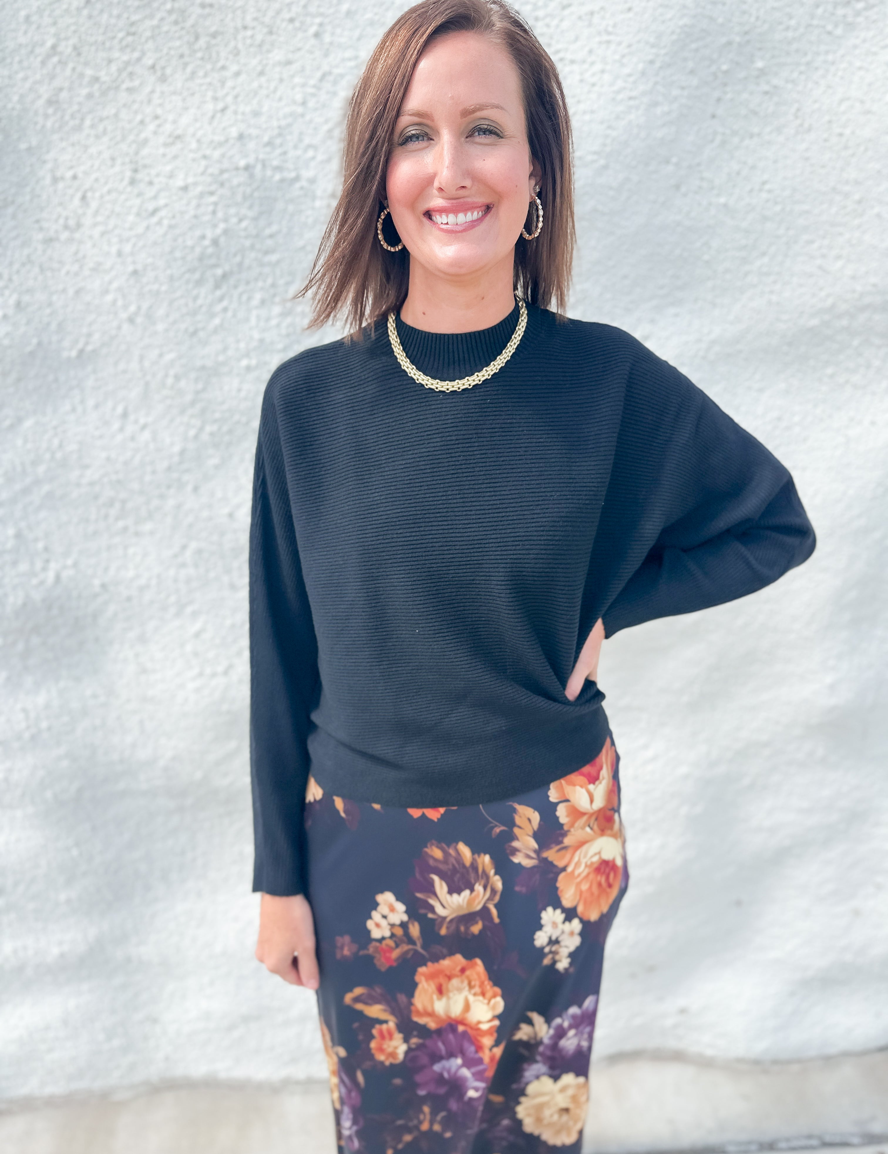 A woman modeling a black ribbed long-sleeve top and a dark floral skirt with orange, purple, and cream flowers, standing against a light gray textured wall.
