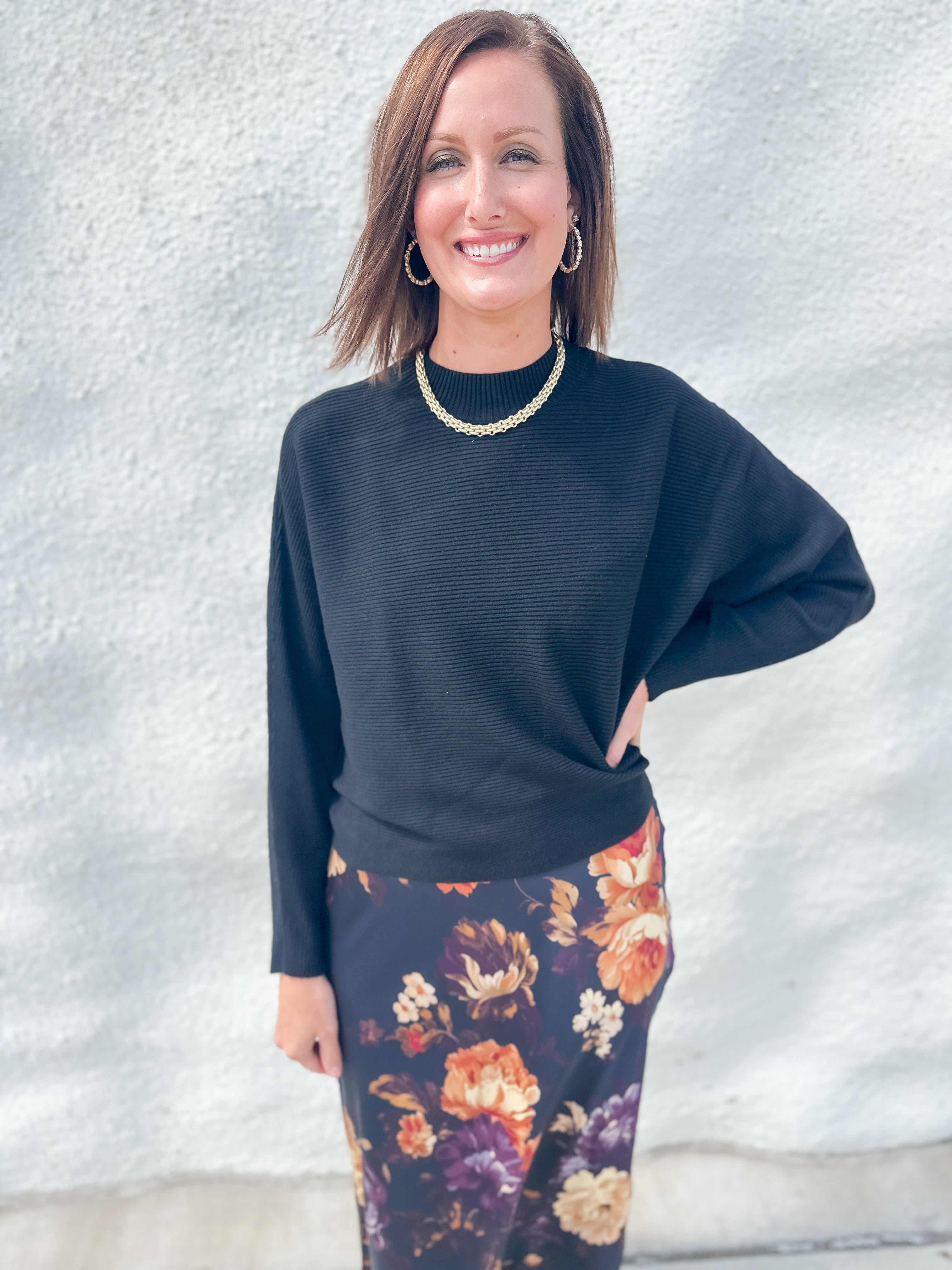 A woman modeling a black ribbed long-sleeve top and a dark floral skirt with orange, purple, and cream flowers, standing against a light gray textured wall.