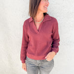A woman wearing a dusty rose ribbed knit polo long sleeve top and gray jeans, standing against a white stucco wall.