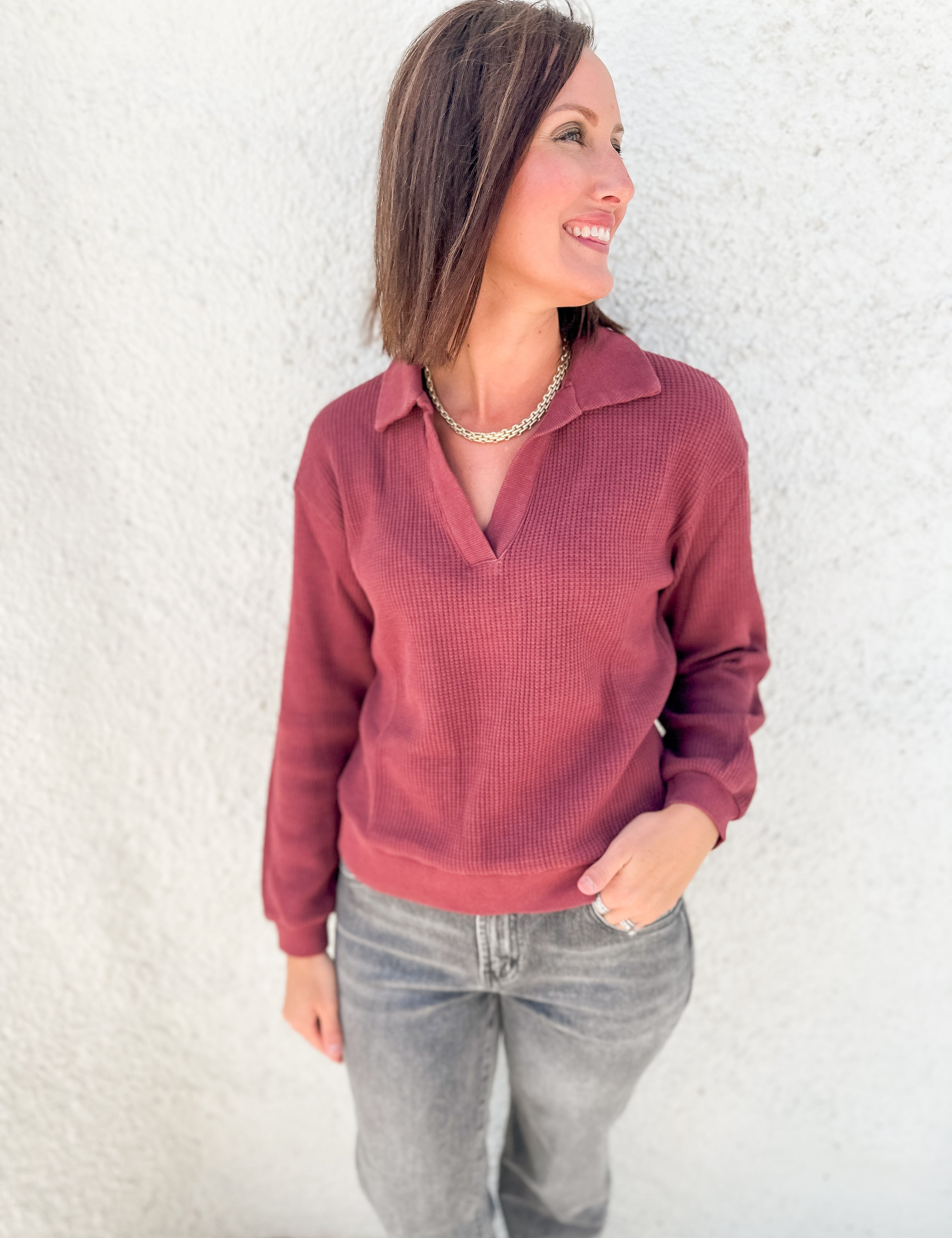 A woman wearing a dusty rose ribbed knit polo long sleeve top and gray jeans, standing against a white stucco wall.