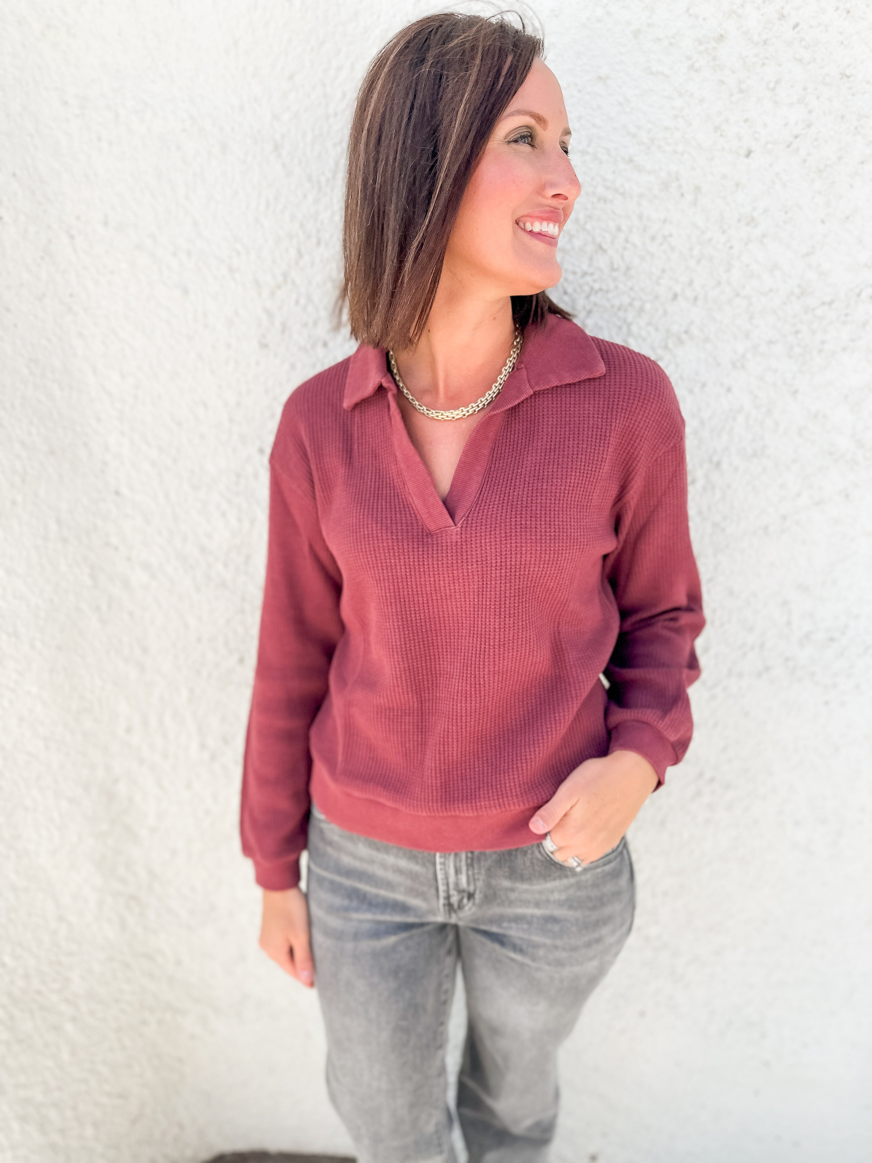 A woman wearing a dusty rose ribbed knit polo long sleeve top and gray jeans, standing against a white stucco wall.