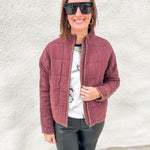 A woman wearing a mahogany quilted zip-up jacket over a white graphic t-shirt and black pants, standing against a white textured wall.
