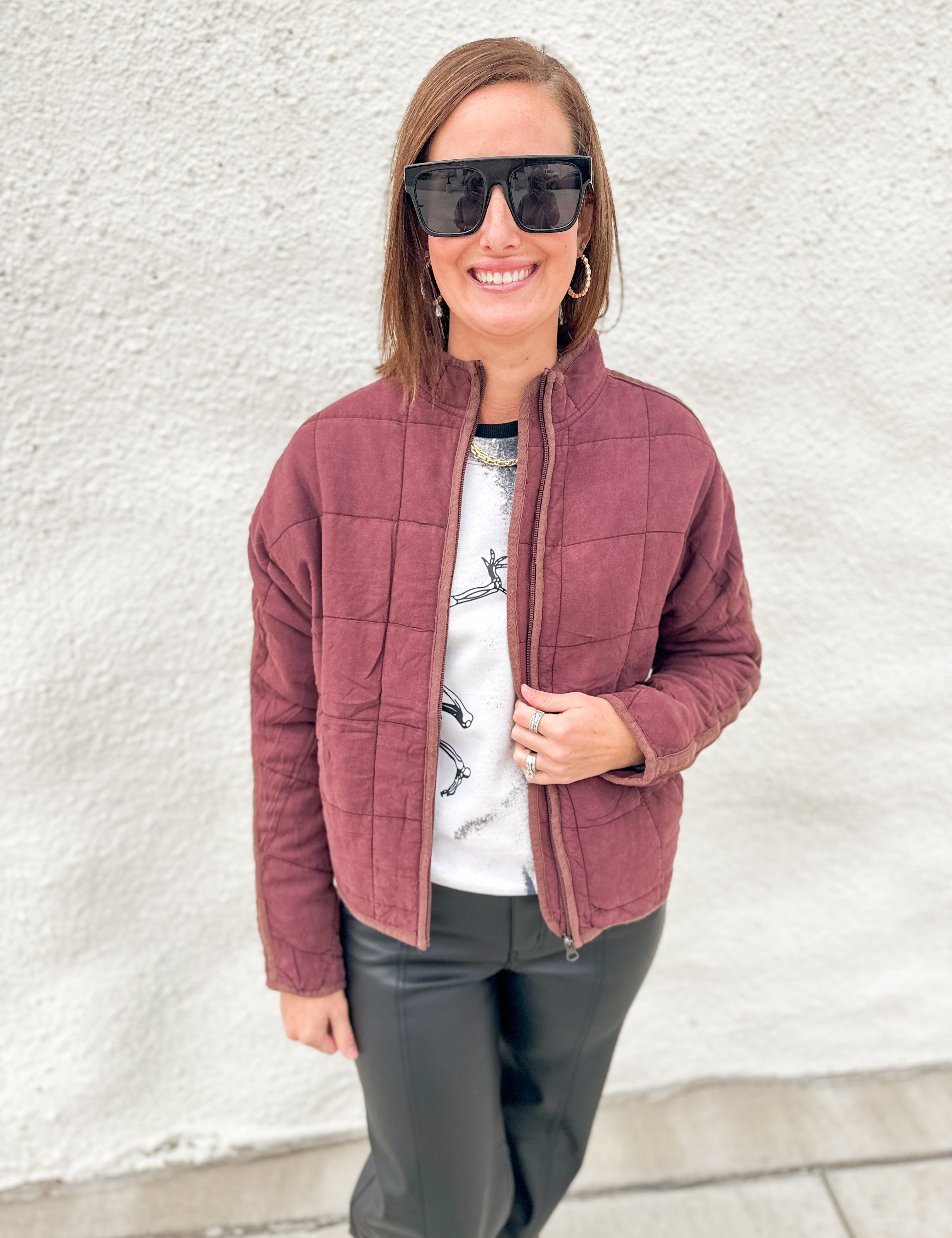 A woman wearing a mahogany quilted zip-up jacket over a white graphic t-shirt and black pants, standing against a white textured wall.