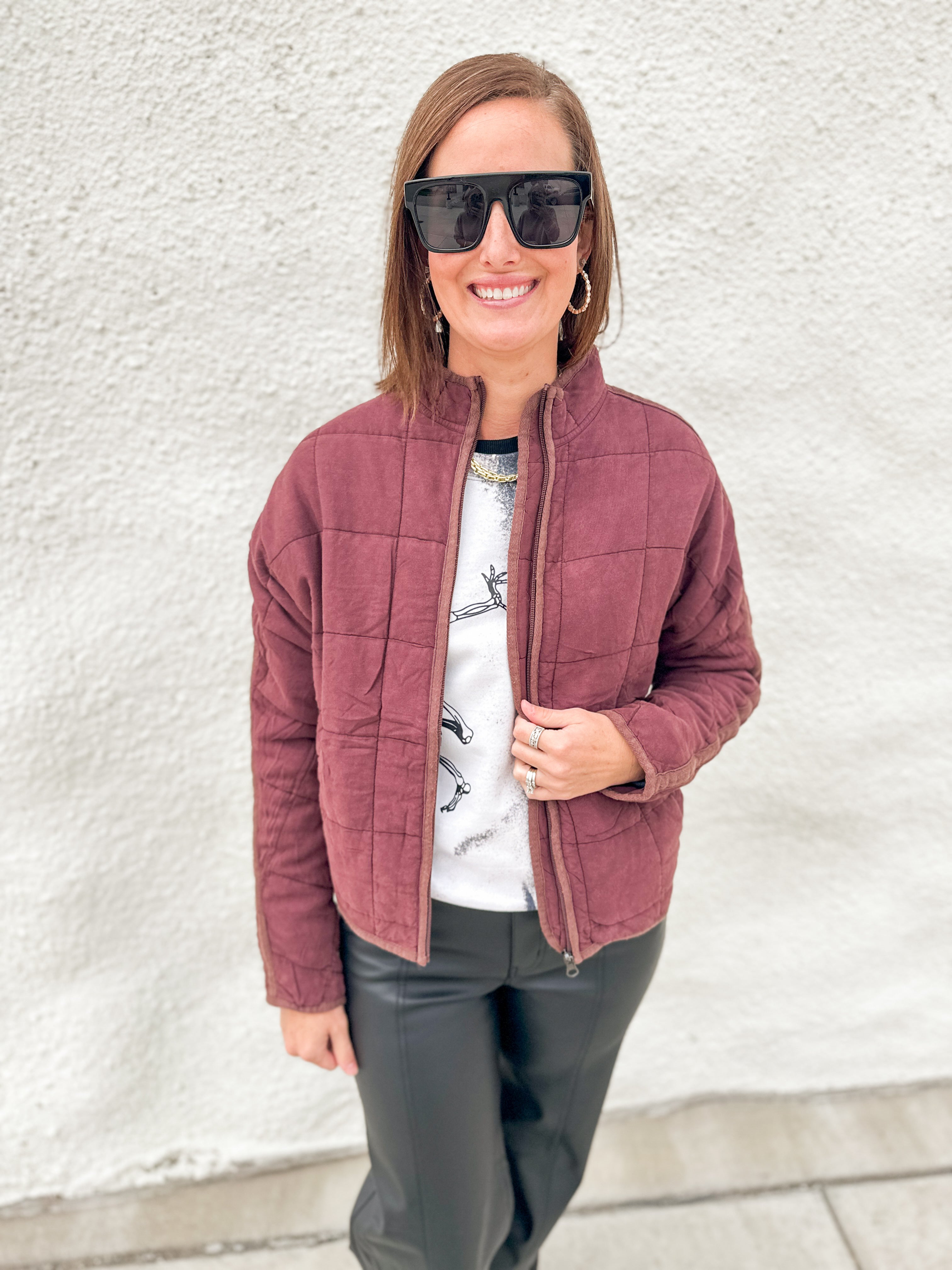 A woman wearing a mahogany quilted zip-up jacket over a white graphic t-shirt and black pants, standing against a white textured wall.