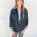 A woman modeling a dark washed denim button-up shirt jacket over a gray ribbed knit top and gray jeans, standing against a white textured wall.