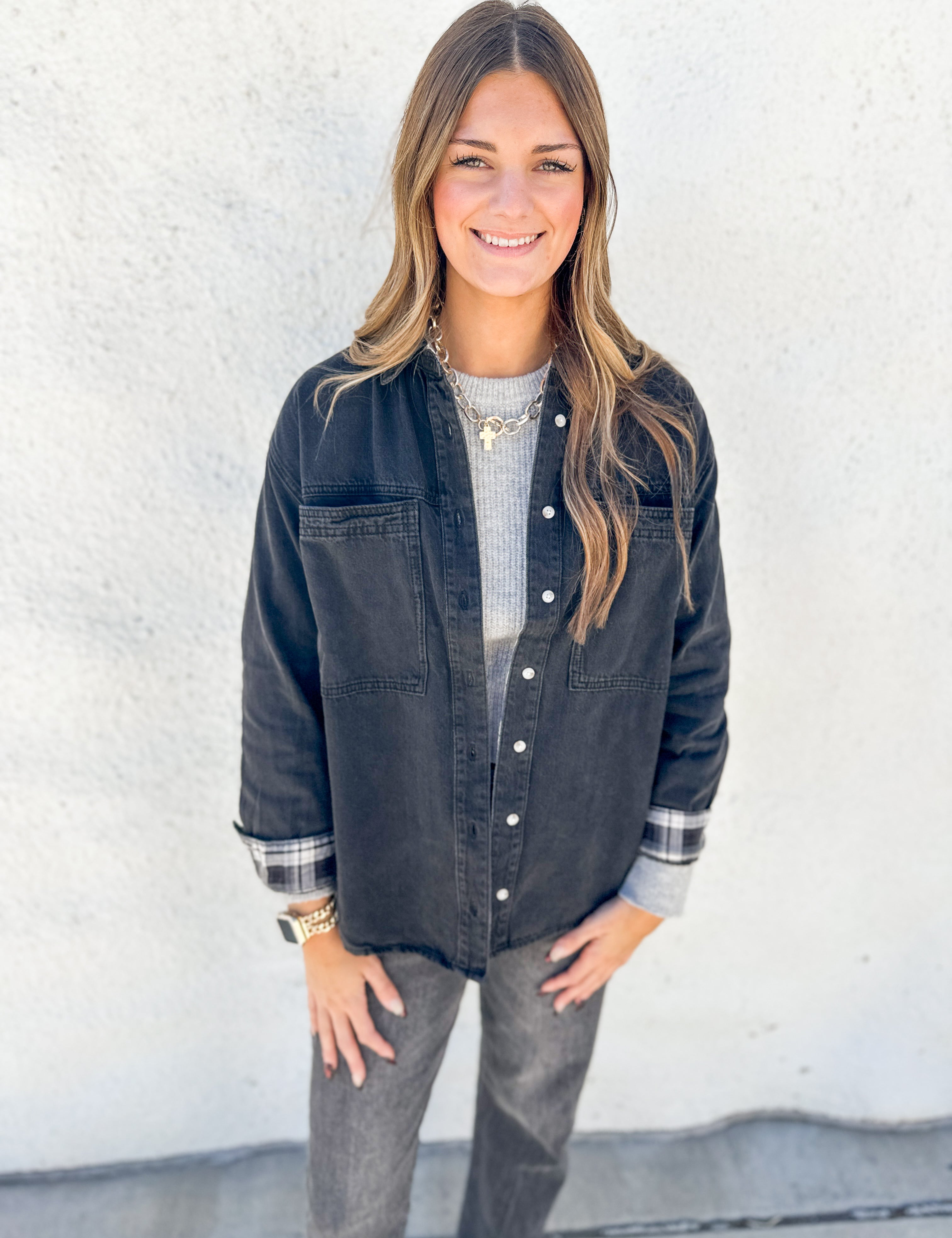 A woman modeling a dark washed denim button-up shirt jacket over a gray ribbed knit top and gray jeans, standing against a white textured wall.