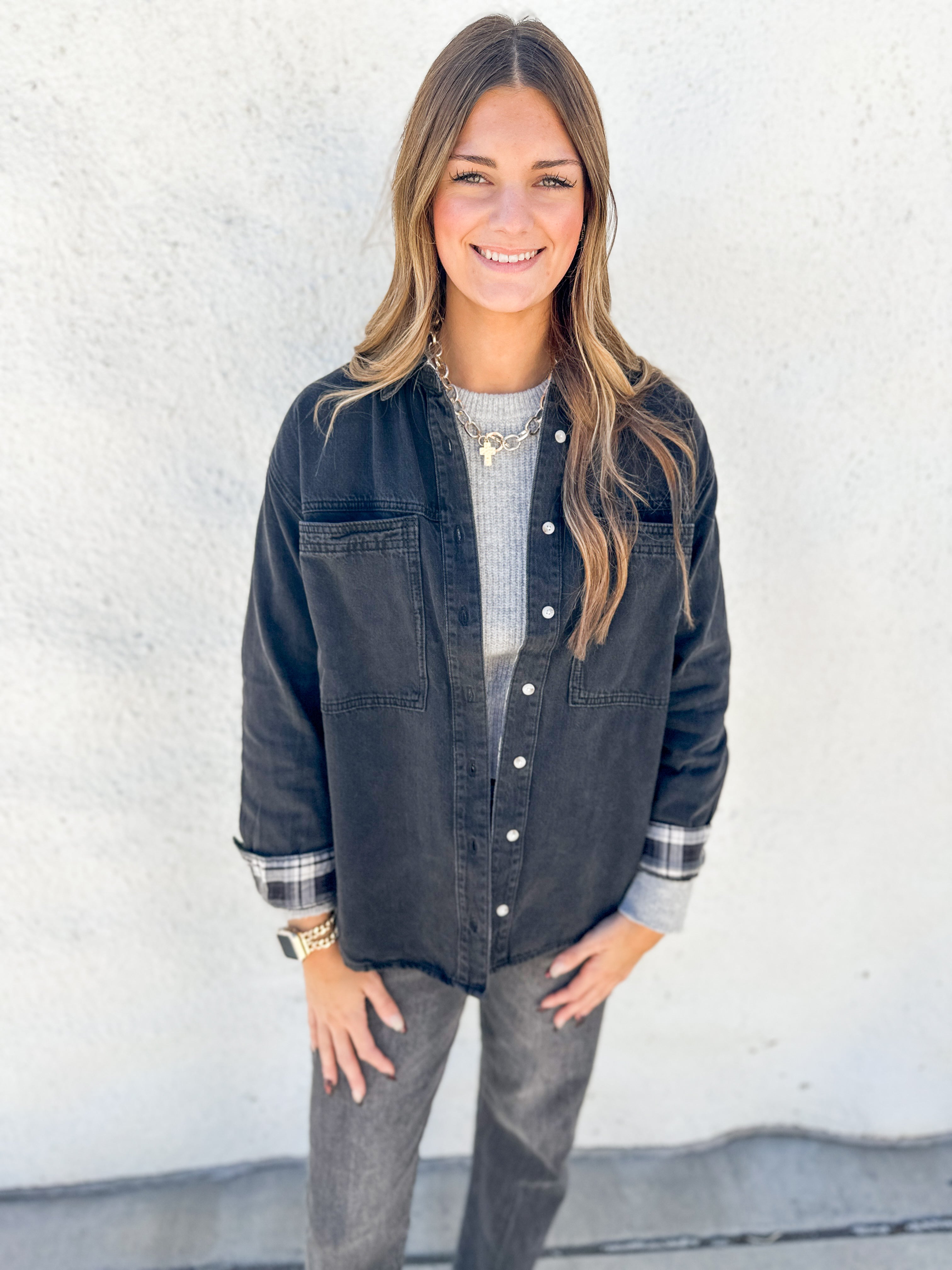 A woman modeling a dark washed denim button-up shirt jacket over a gray ribbed knit top and gray jeans, standing against a white textured wall.