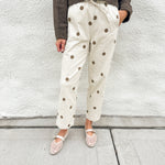 A person wearing cream-colored barrel pants with brown polka dots and elastic drawstring waist, a dark brown long-sleeve top, and cream mesh mary jane flats, standing against a white textured wall.