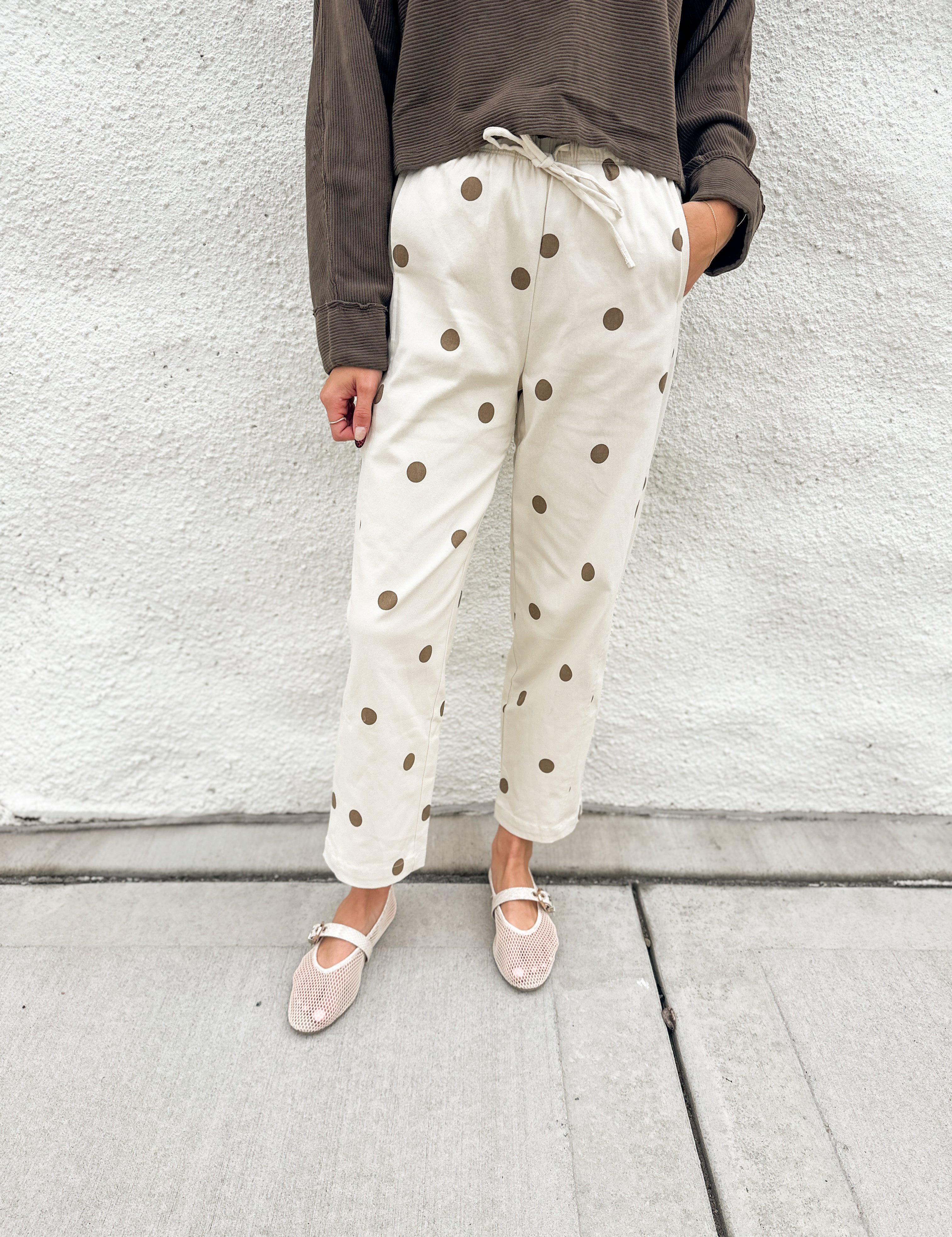 A person wearing cream-colored barrel pants with brown polka dots and elastic drawstring waist, a dark brown long-sleeve top, and cream mesh mary jane flats, standing against a white textured wall.