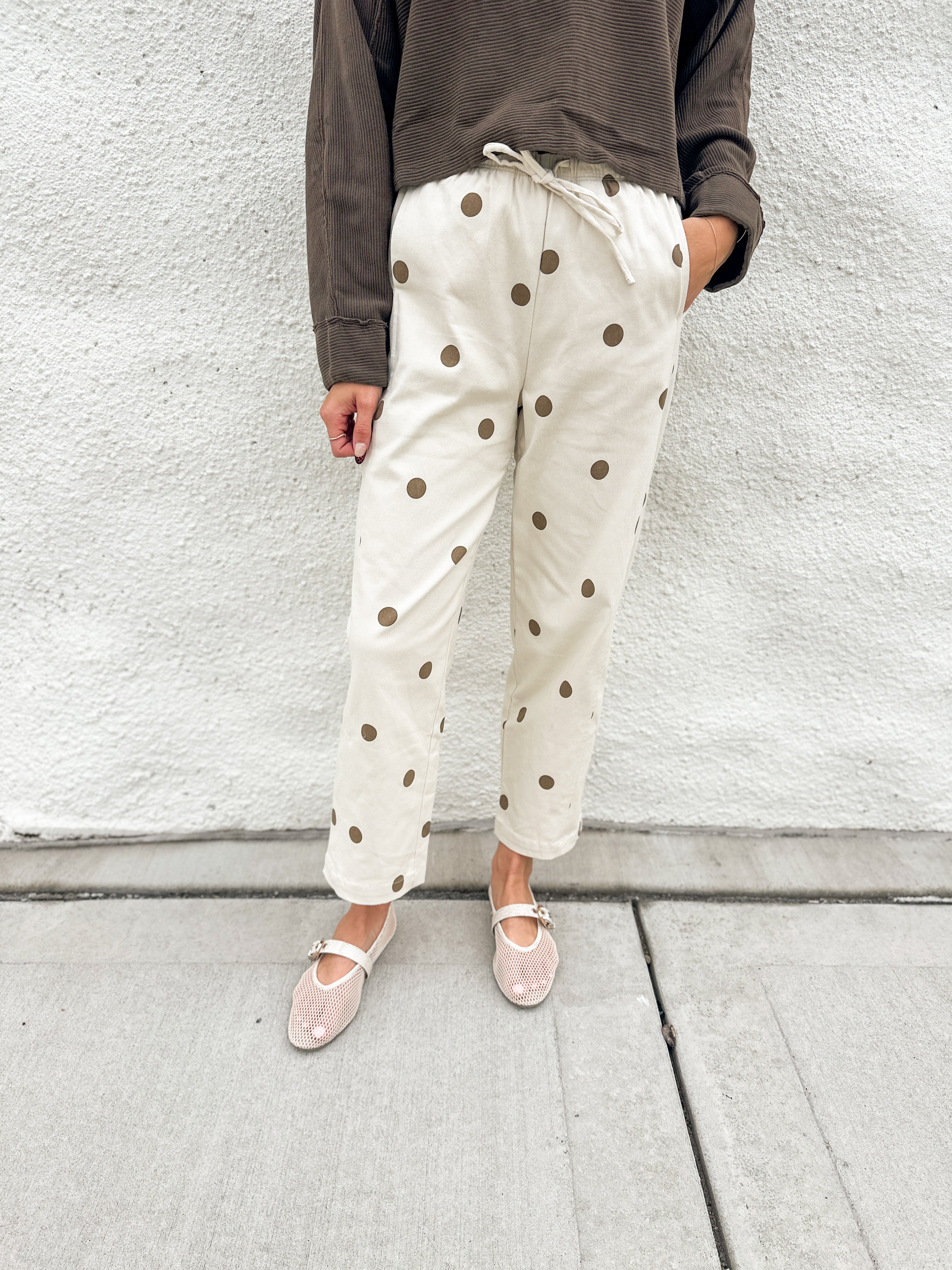 A person wearing cream-colored barrel pants with brown polka dots and elastic drawstring waist, a dark brown long-sleeve top, and cream mesh mary jane flats, standing against a white textured wall.