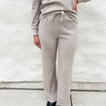A person wearing a light taupe heathered lounge set consisting of a long-sleeve top and matching wide-leg sweatpants, standing against a white textured wall.
