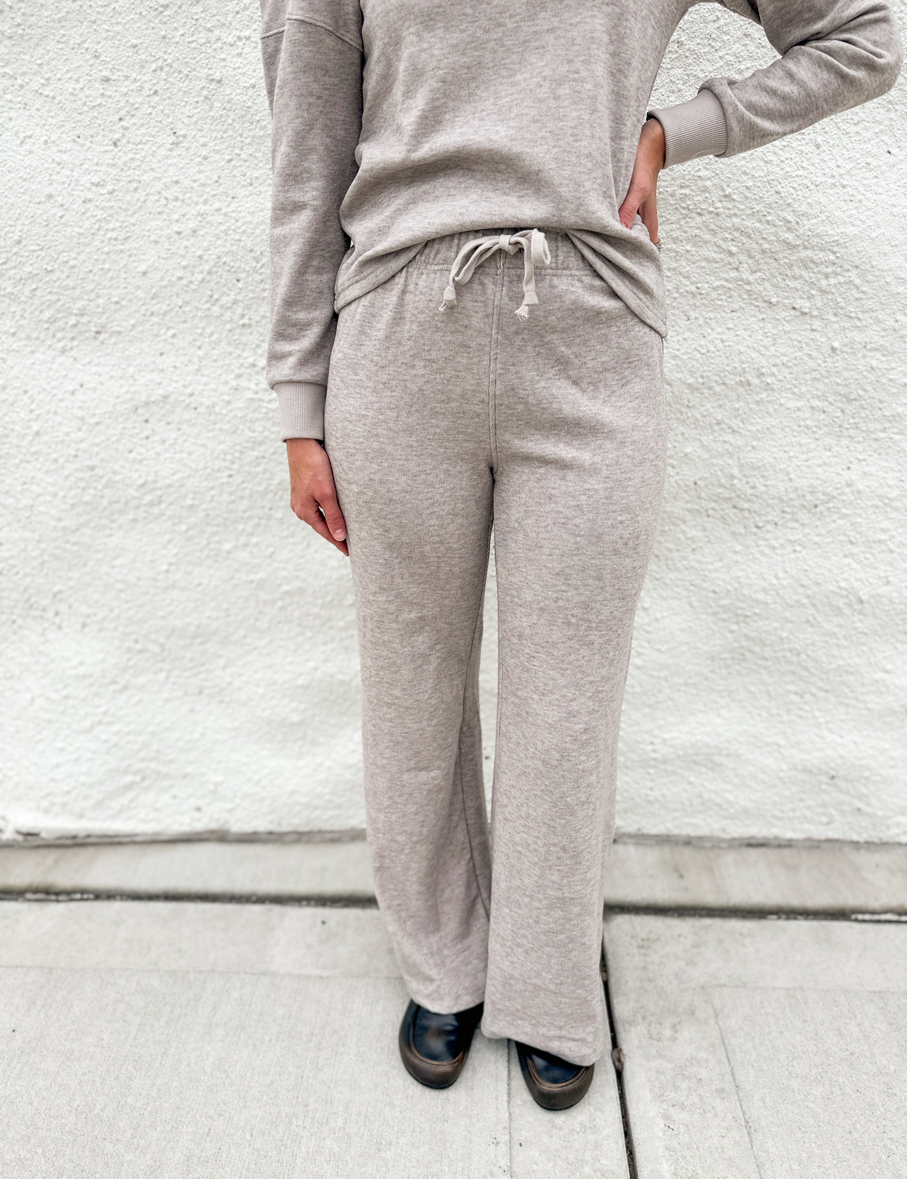 A person wearing a light taupe heathered lounge set consisting of a long-sleeve top and matching wide-leg sweatpants, standing against a white textured wall.