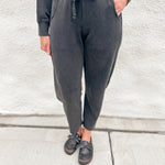 A person wearing dark charcoal gray jogger barrel sweatpants with a drawstring waist, paired with a matching dark sweatshirt and black loafer clogs, standing against a light gray textured wall.