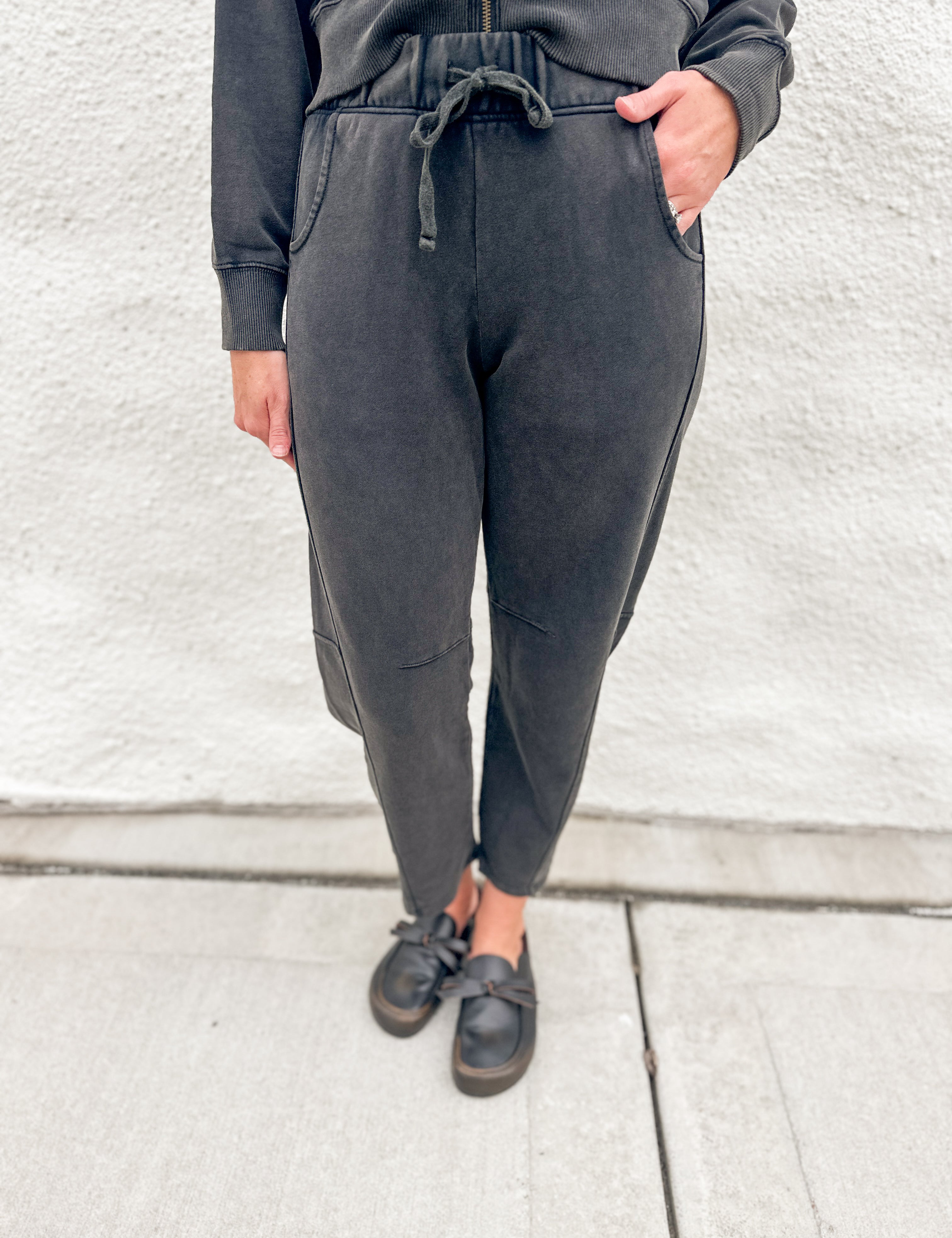 A person wearing dark charcoal gray jogger barrel sweatpants with a drawstring waist, paired with a matching dark sweatshirt and black loafer clogs, standing against a light gray textured wall.