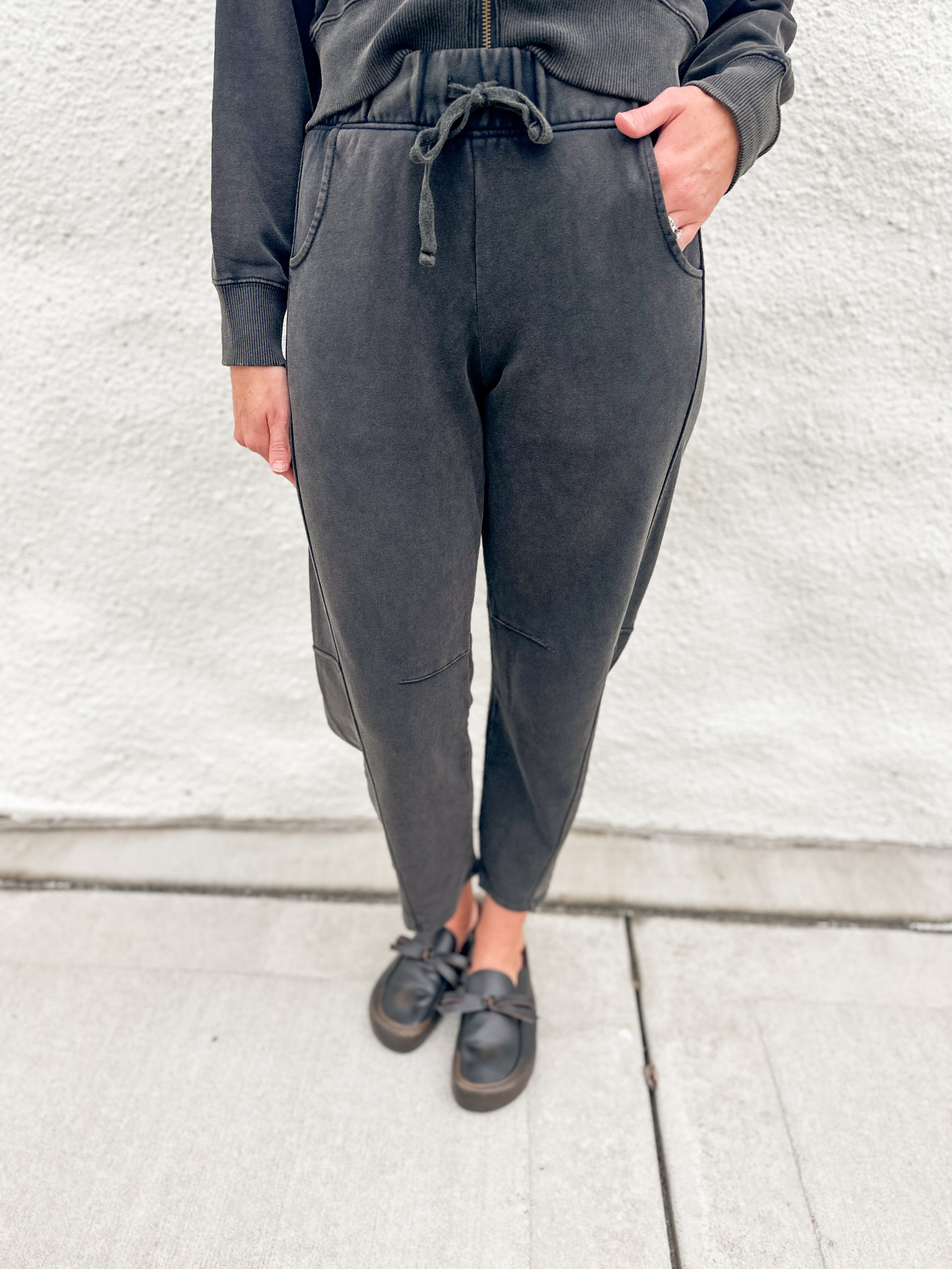 A person wearing dark charcoal gray jogger barrel sweatpants with a drawstring waist, paired with a matching dark sweatshirt and black loafer clogs, standing against a light gray textured wall.