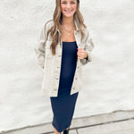 A woman stands against a white textured wall wearing a light grey denim jacket over a dark navy midi dress, paired with black ankle boots and layered gold necklaces.