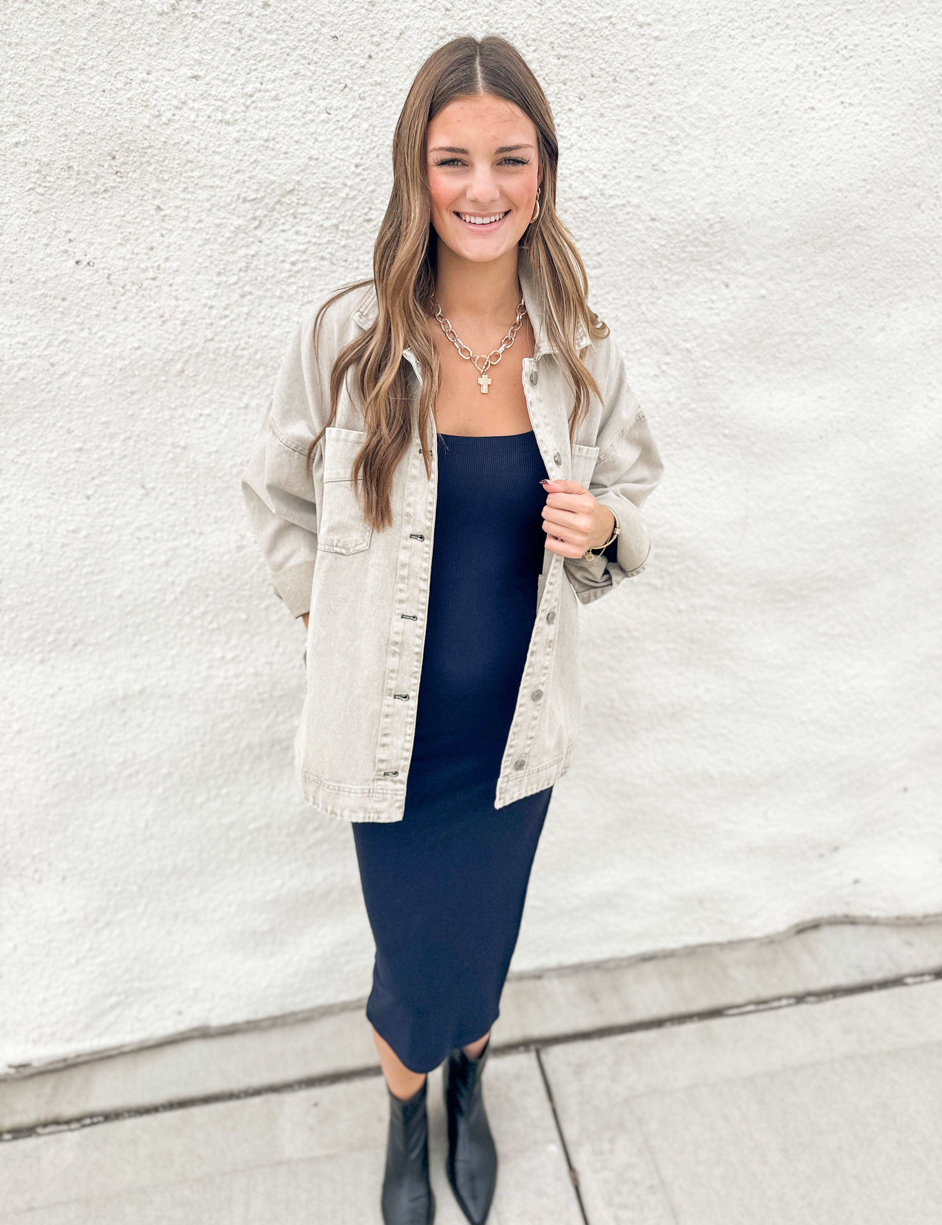 A woman stands against a white textured wall wearing a light grey denim jacket over a dark navy midi dress, paired with black ankle boots and layered gold necklaces.