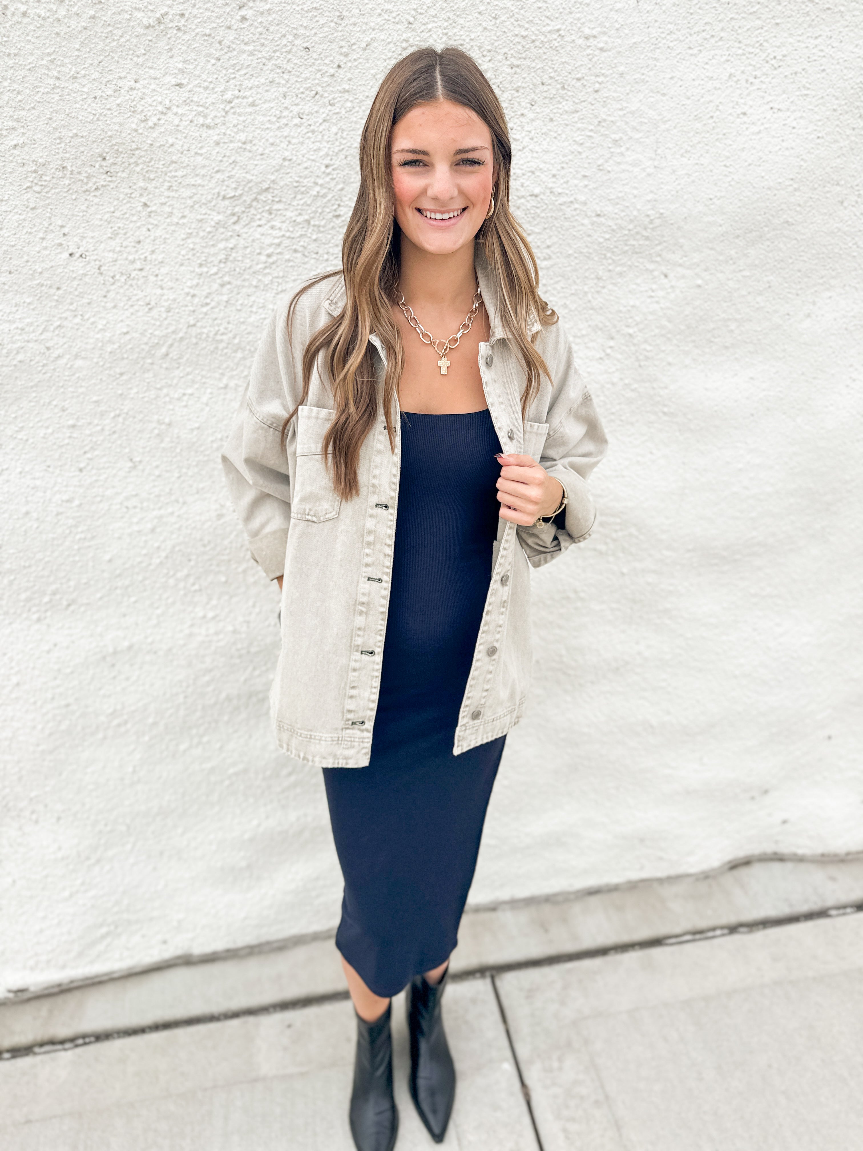 A woman stands against a white textured wall wearing a light grey denim jacket over a dark navy midi dress, paired with black ankle boots and layered gold necklaces.