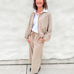 A woman stands outdoors against a light gray stucco wall wearing a beige zip-front bubble hem sweater bomber with suede accents, matching wide-leg pants, a white collared shirt, and black shoes.