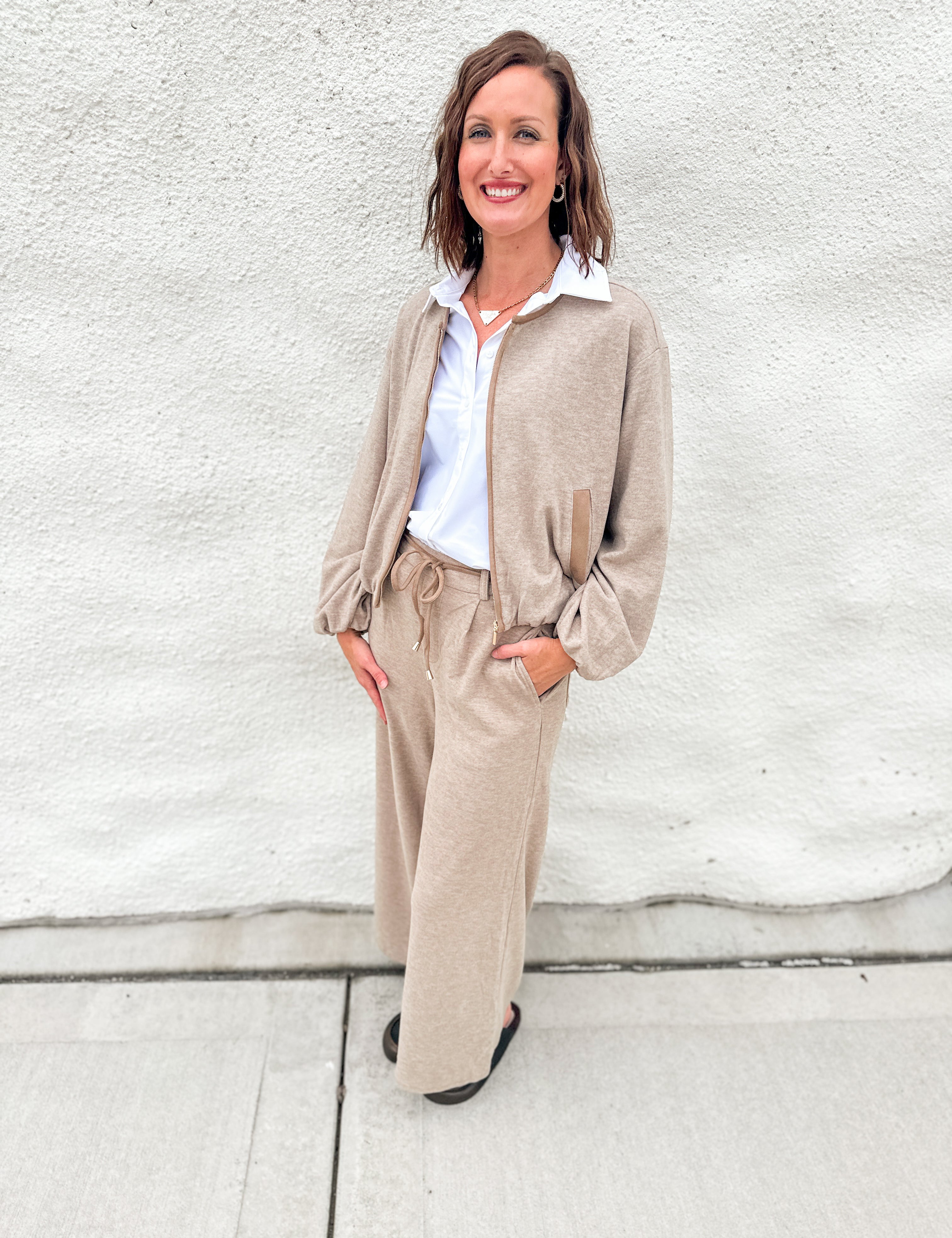 A woman stands outdoors against a light gray stucco wall wearing a beige zip-front bubble hem sweater bomber with suede accents, matching wide-leg pants, a white collared shirt, and black shoes.