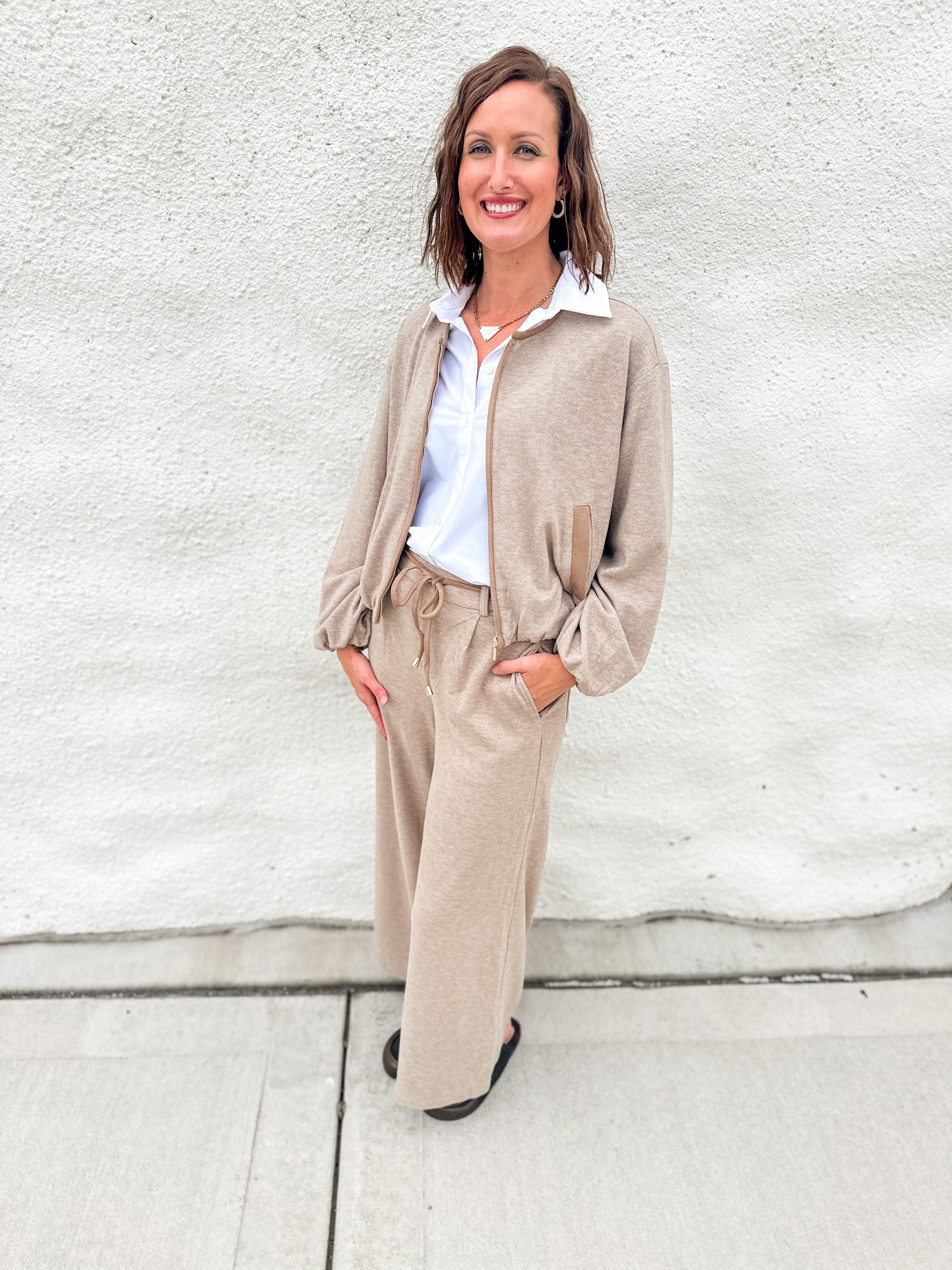 A woman stands outdoors against a light gray stucco wall wearing a beige zip-front bubble hem sweater bomber with suede accents, matching wide-leg pants, a white collared shirt, and black shoes.