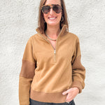A woman wearing a tan quarter-zip pullover sweater with ribbed trim and darker brown ribbed elbow patches, paired with dark gray jeans, standing against a textured white wall.