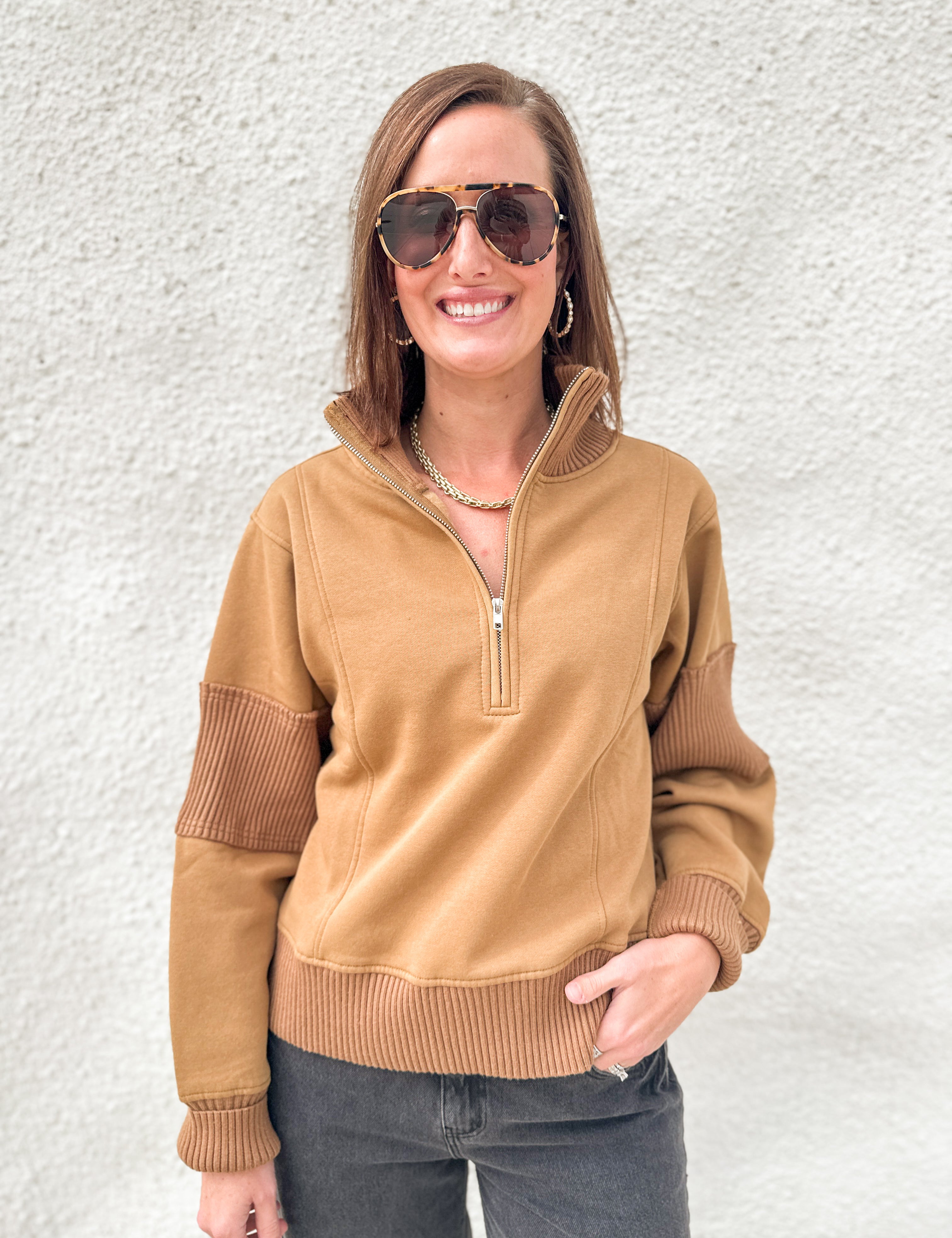 A woman wearing a tan quarter-zip pullover sweater with ribbed trim and darker brown ribbed elbow patches, paired with dark gray jeans, standing against a textured white wall.
